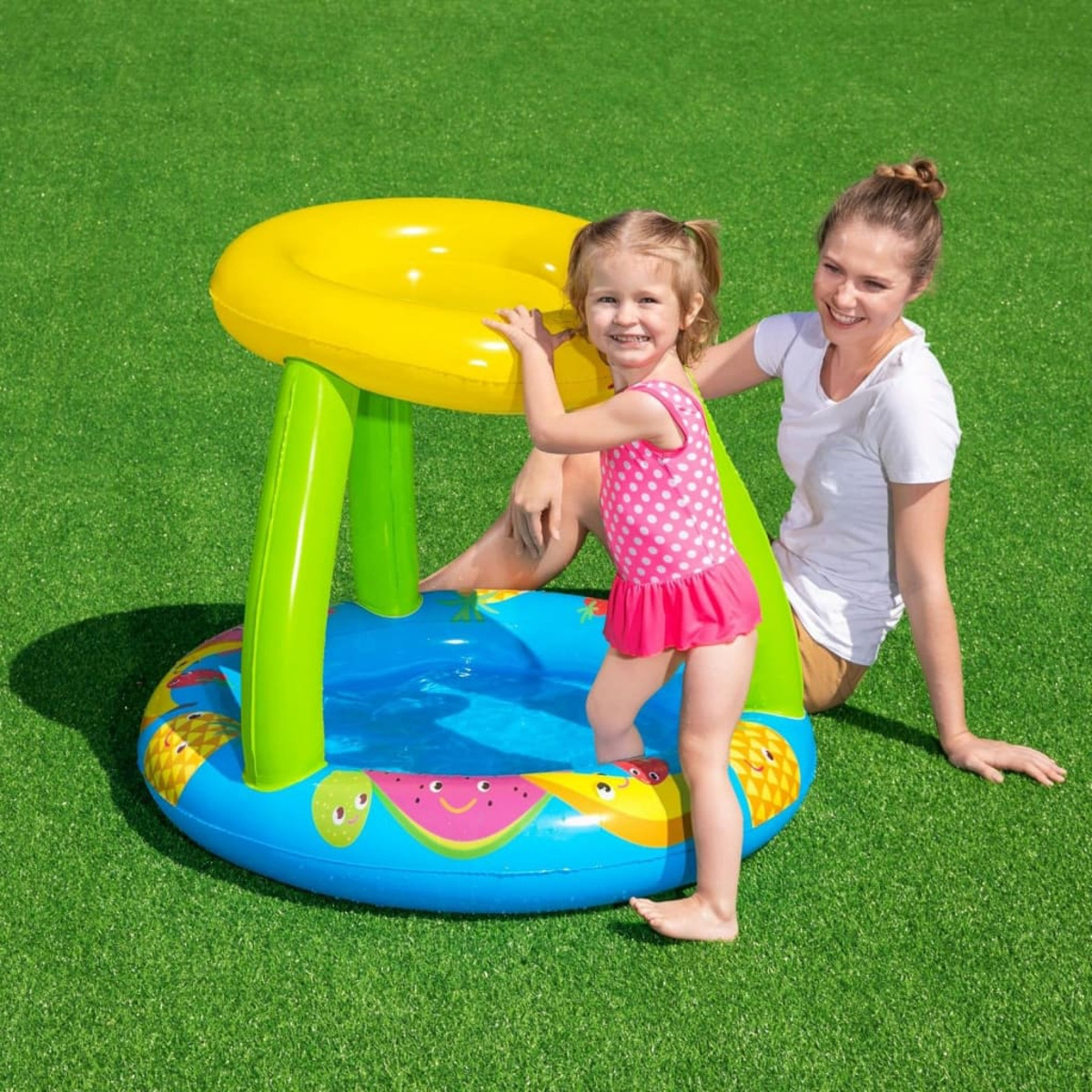 Ein Mädchen in einem rosa Badeanzug steht in einem blauen Pool unter einem gelben Vordach, eine Frau sitzt in der Nähe auf Gras.