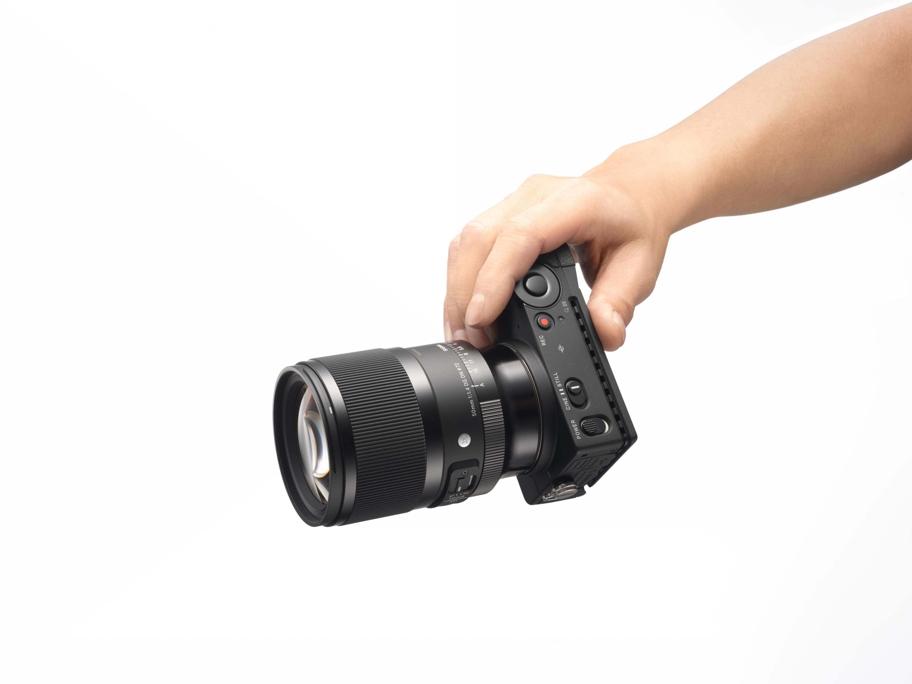 Een hand die een zwarte camera met een lens vasthoudt. De hand is het enige element op de voorgrond. De achtergrond is wit.