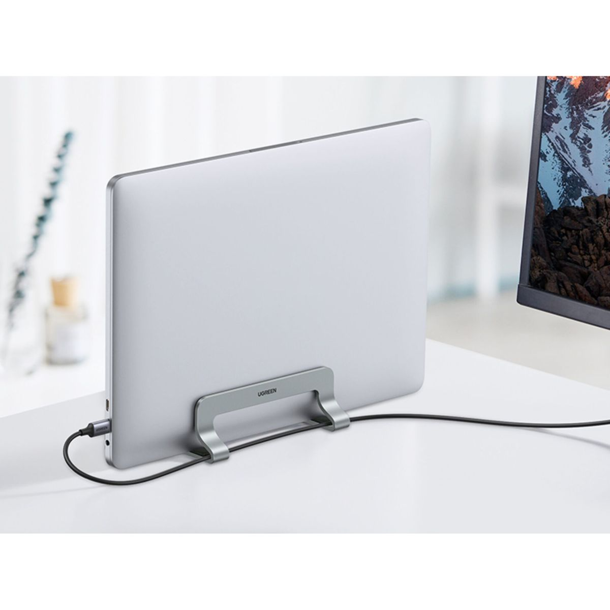 Ein Laptop in einem silbernen Ständer mit angeschlossenem Kabel, ein Monitor rechts, weiße Oberfläche.