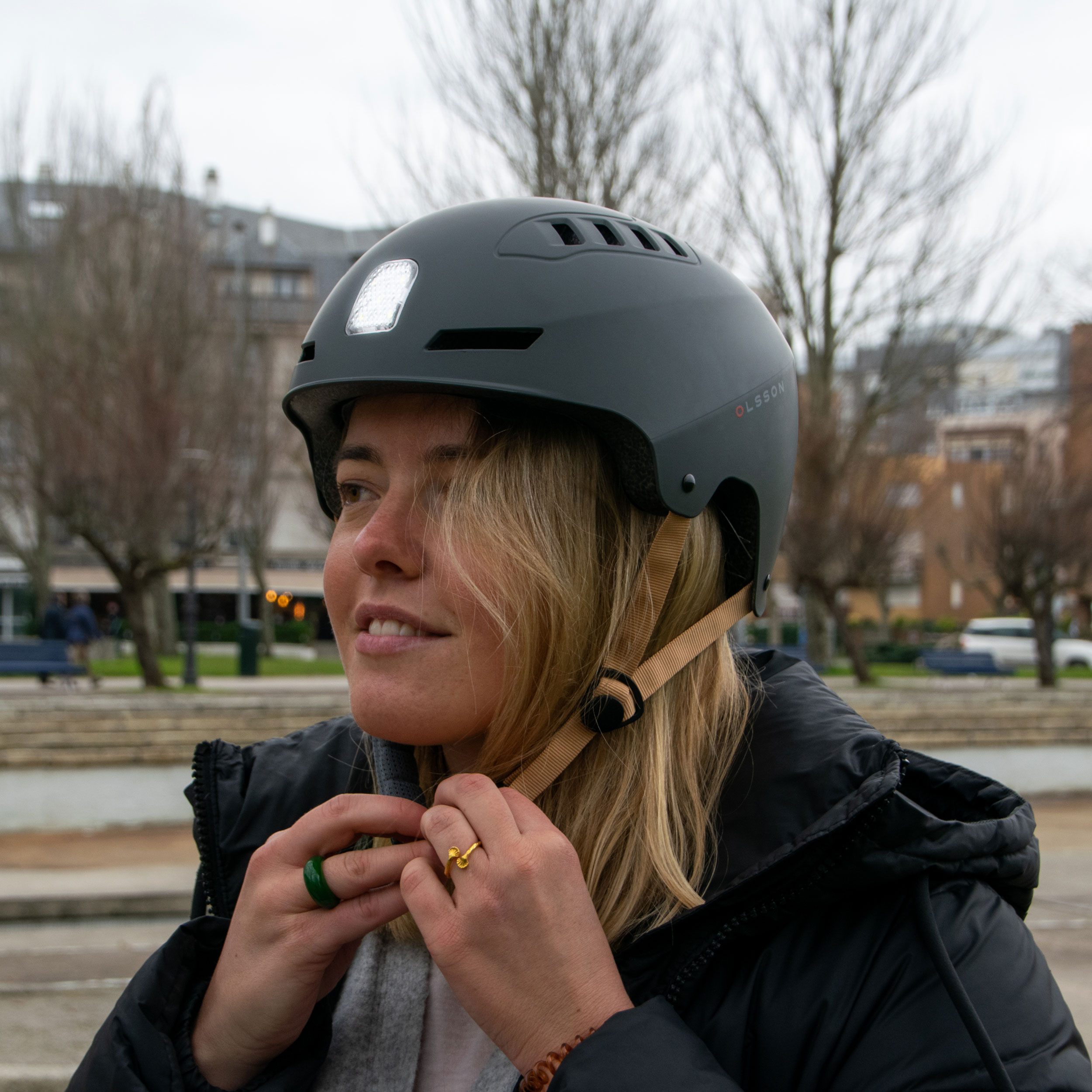 Una mujer con casco gris con luz, sonriendo y ajustando la correa. Los árboles están al fondo.