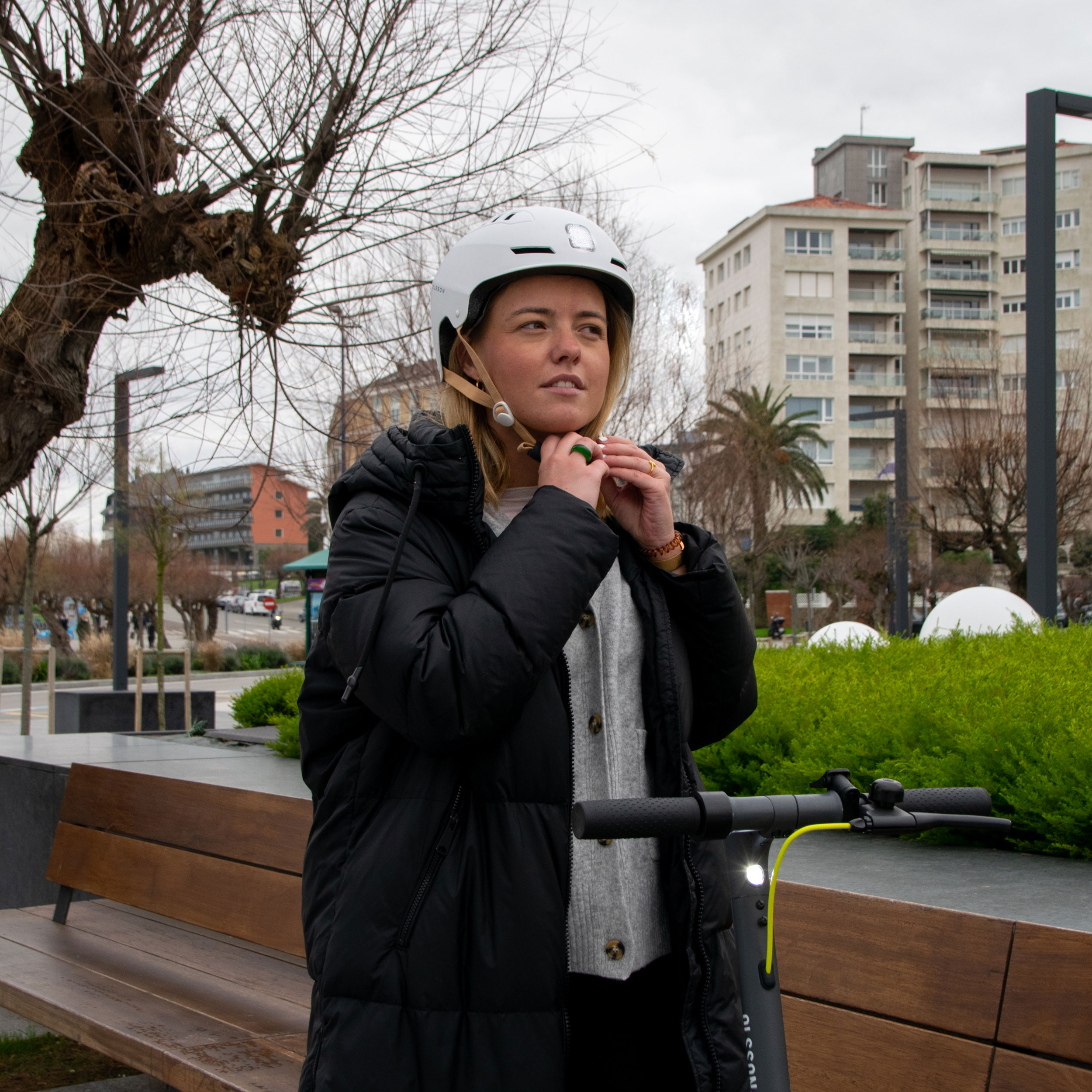 Mujer ajustando un casco blanco. Lleva un abrigo negro y está cerca de un scooter.