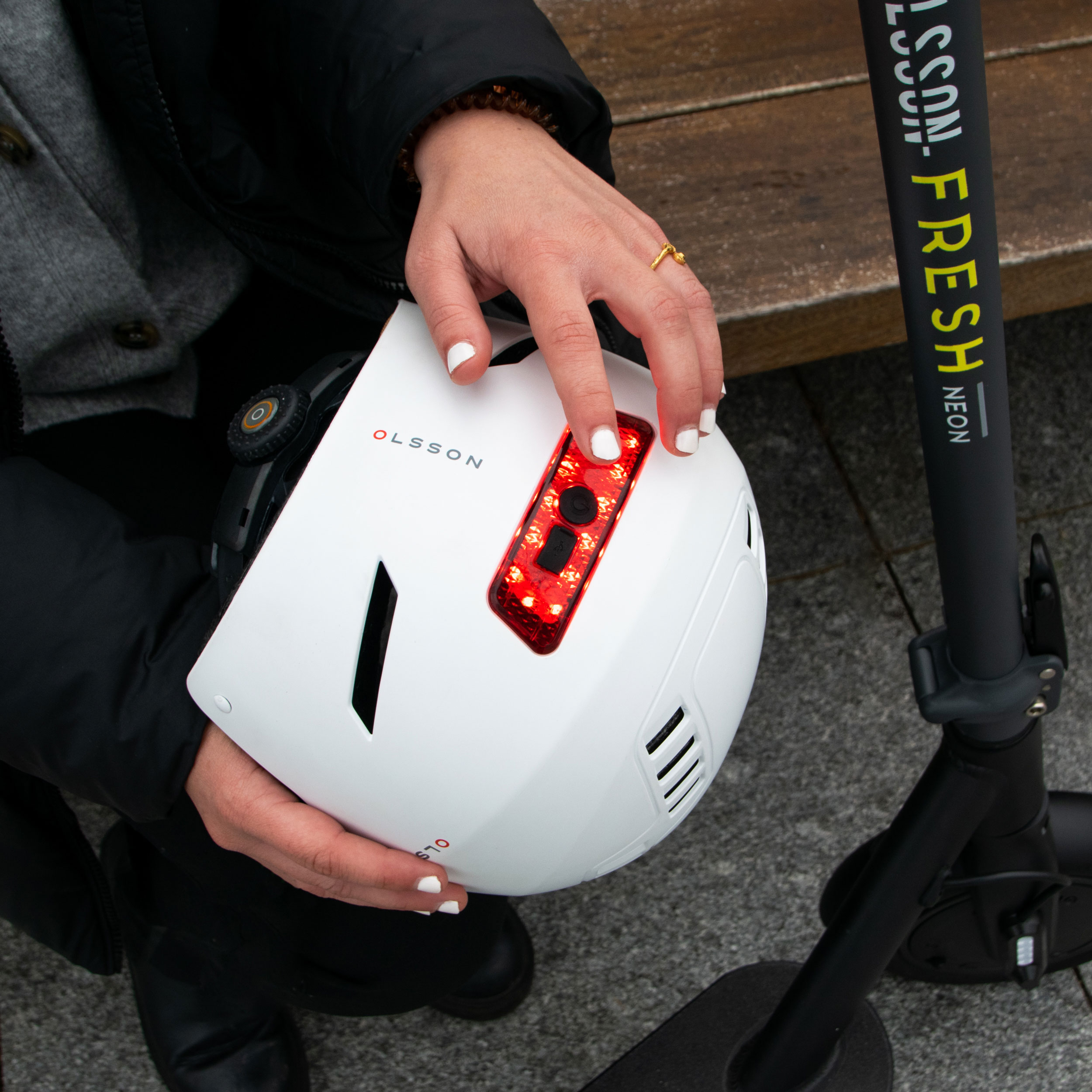 Una mano presiona una luz roja en un casco Olsson blanco junto a un patinete negro.