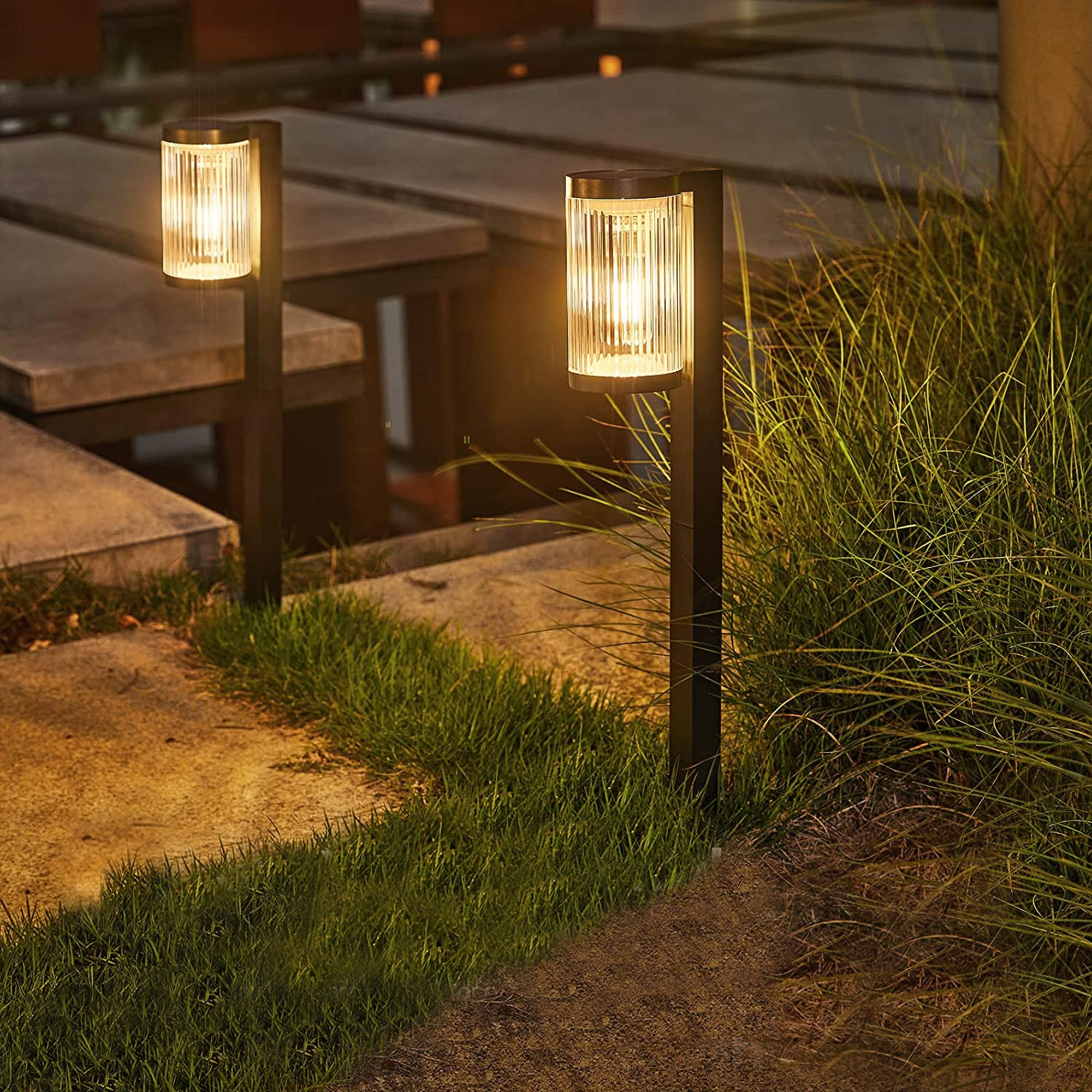 Zwei beleuchtete Außenleuchten auf Pfosten neben einem grasbewachsenen Weg. Hintergrund umfasst Steinsitzplätze und weitere Lichter.