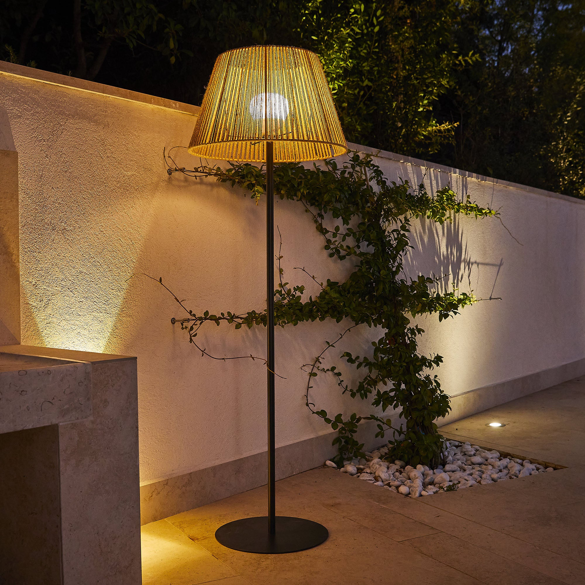 Außenlampe mit gewebtem Schirm, beleuchtet. Neben einer weißen Wand mit Grün und Steingrund.