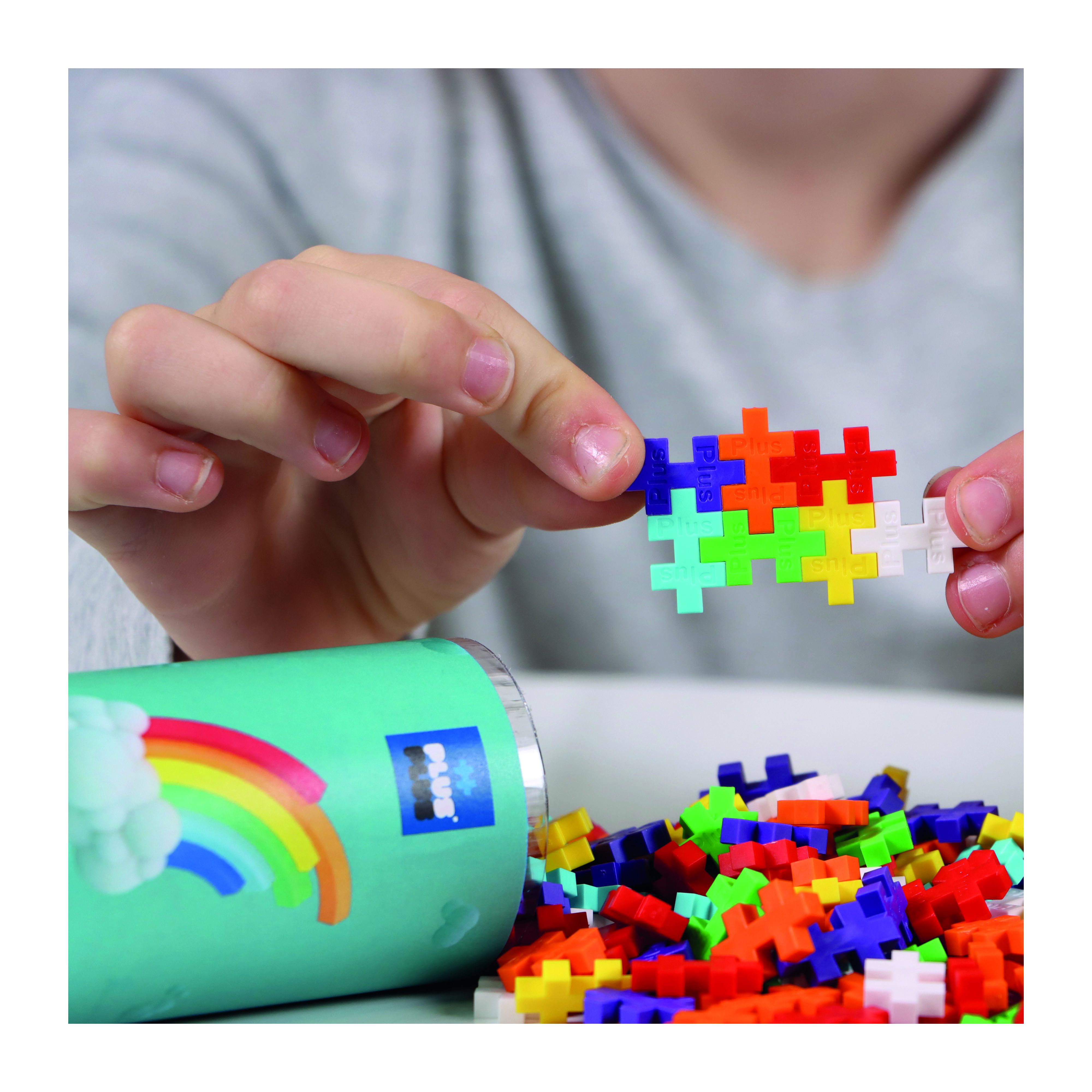 Hände halten bunte Kunststoff-Spielzeugblöcke; ein blauer Behälter mit Regenbogen-Logo daneben.