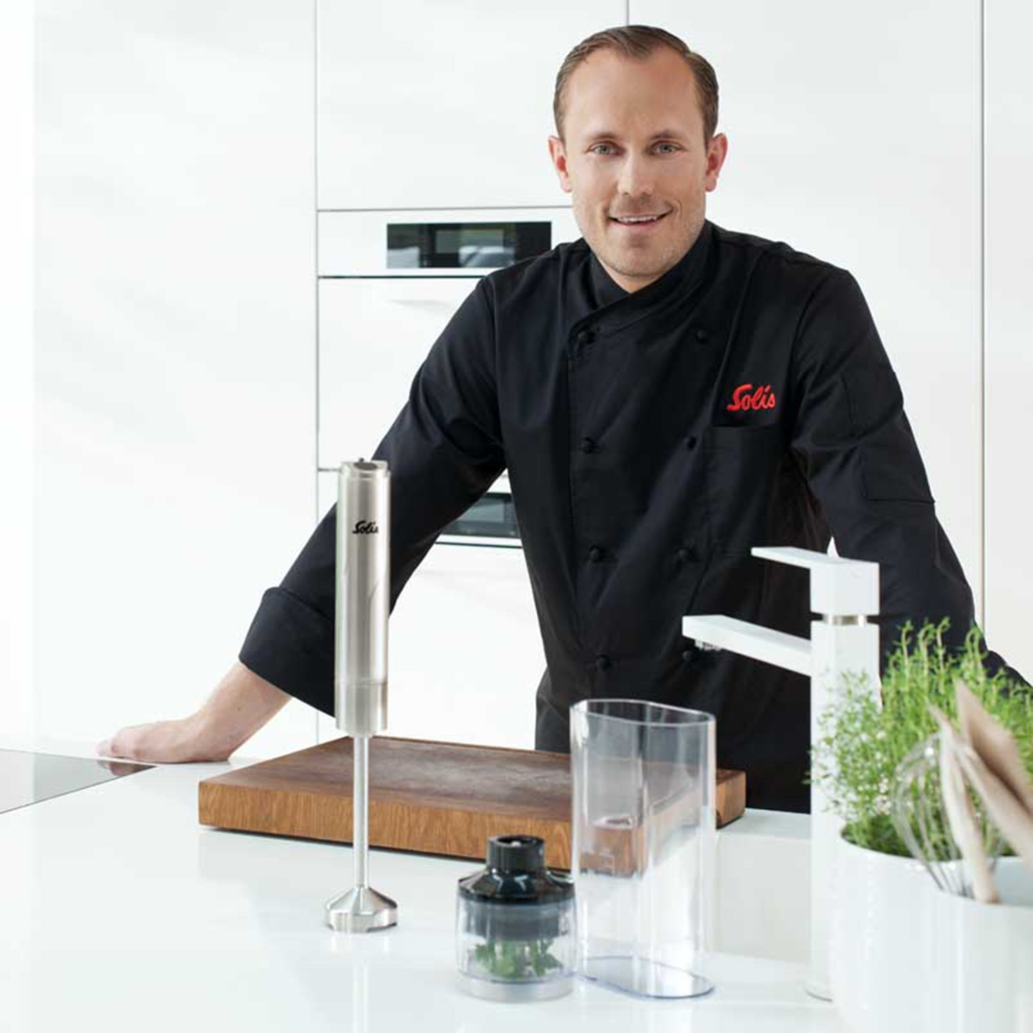 Uno chef in uniforme nera con un frullatore Solis, tagliere in legno e un contenitore trasparente.