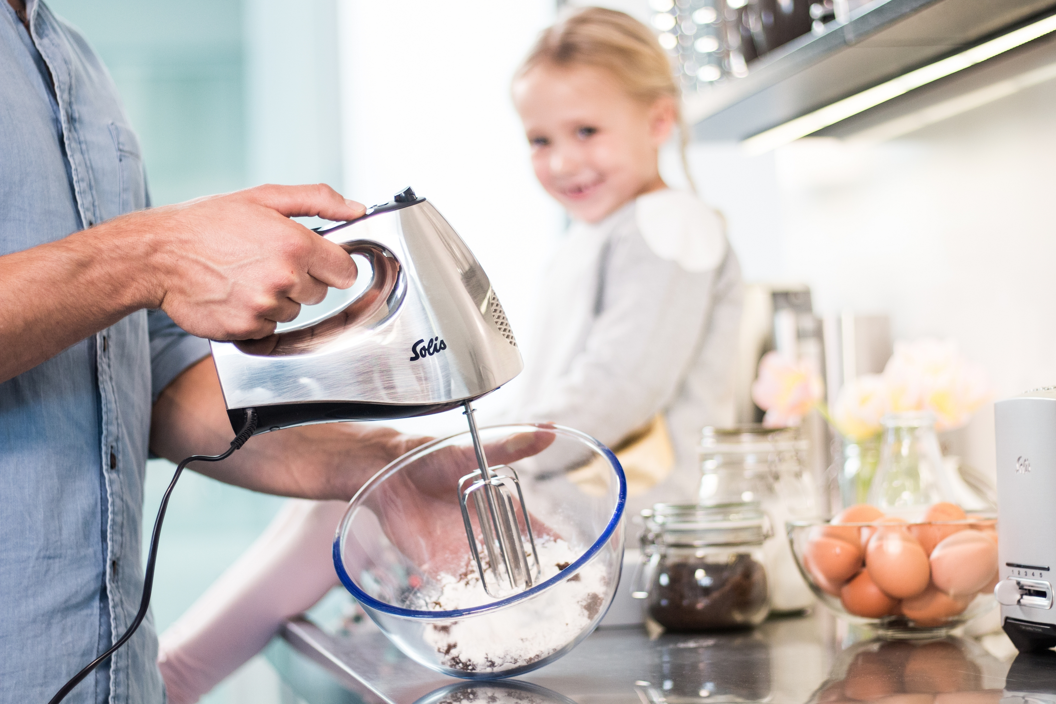 Eine Person benutzt einen silbernen Handmixer, um Zutaten in einer Glasschüssel zu verrühren, ein Kind schaut zu.