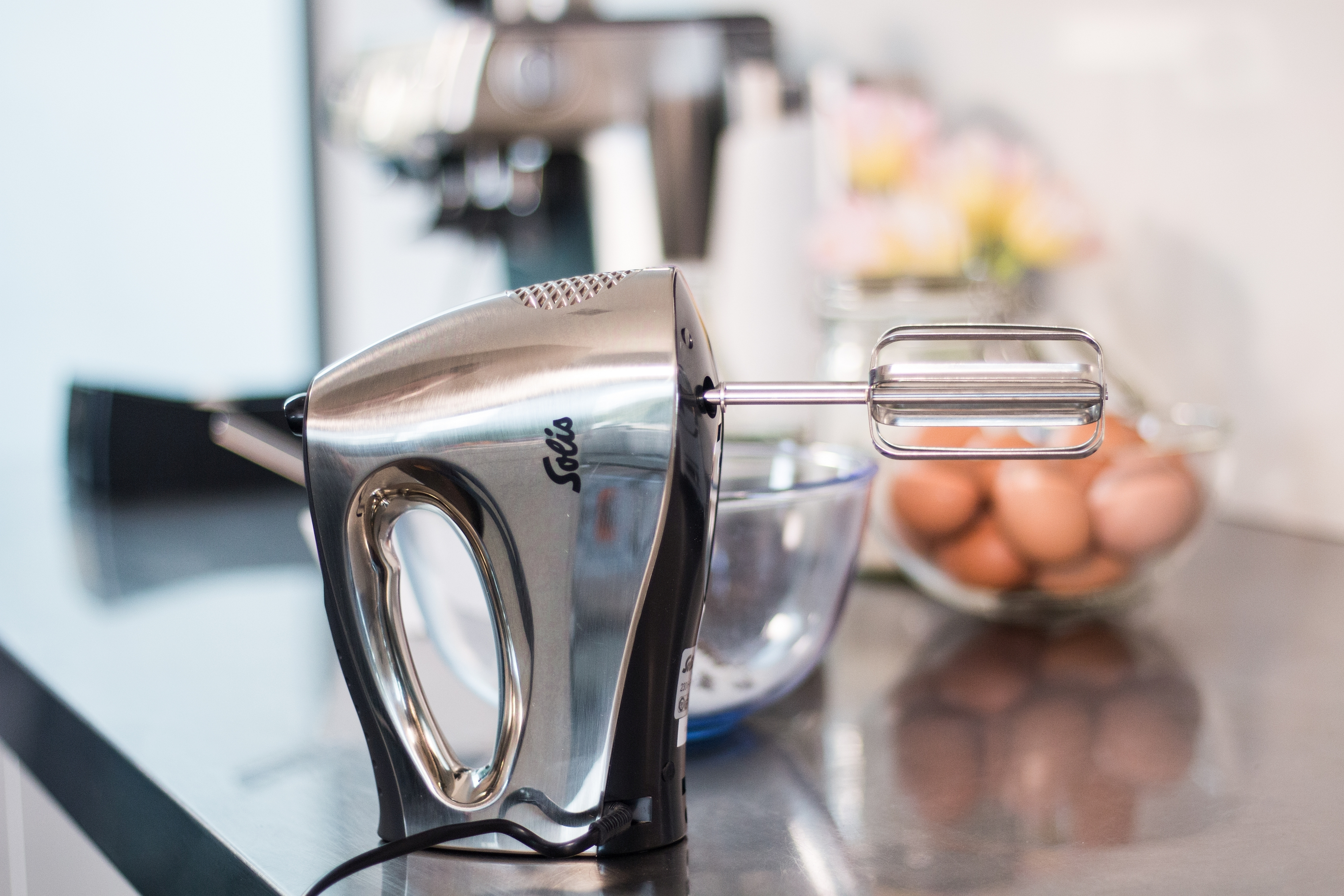Silberner Handmixer auf Edelstahl-Arbeitsplatte. Eier und eine Rührschüssel im Hintergrund.