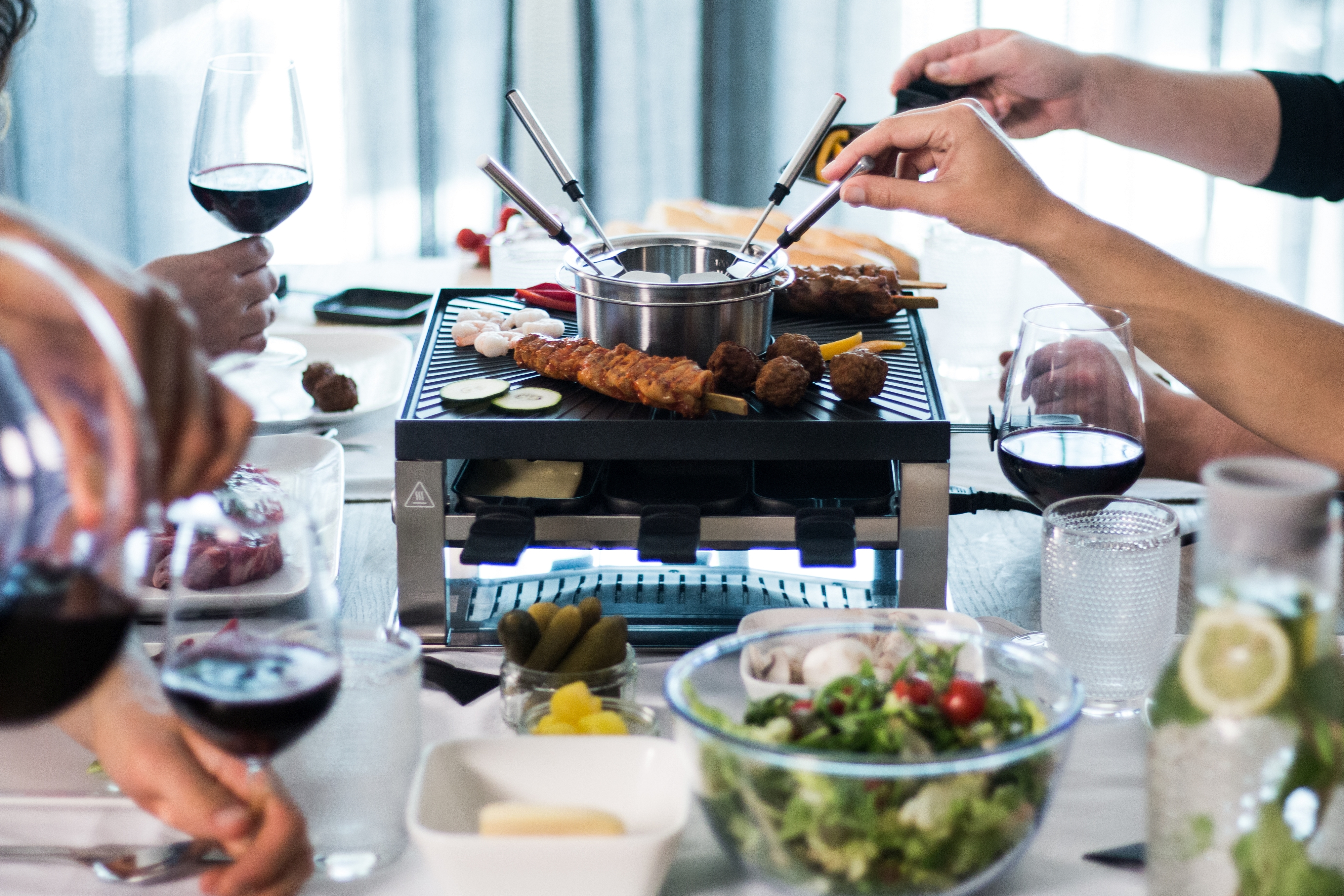 Ludzie przy stole z jedzeniem, garnkiem do fondue, kieliszkami do wina i grillem.
