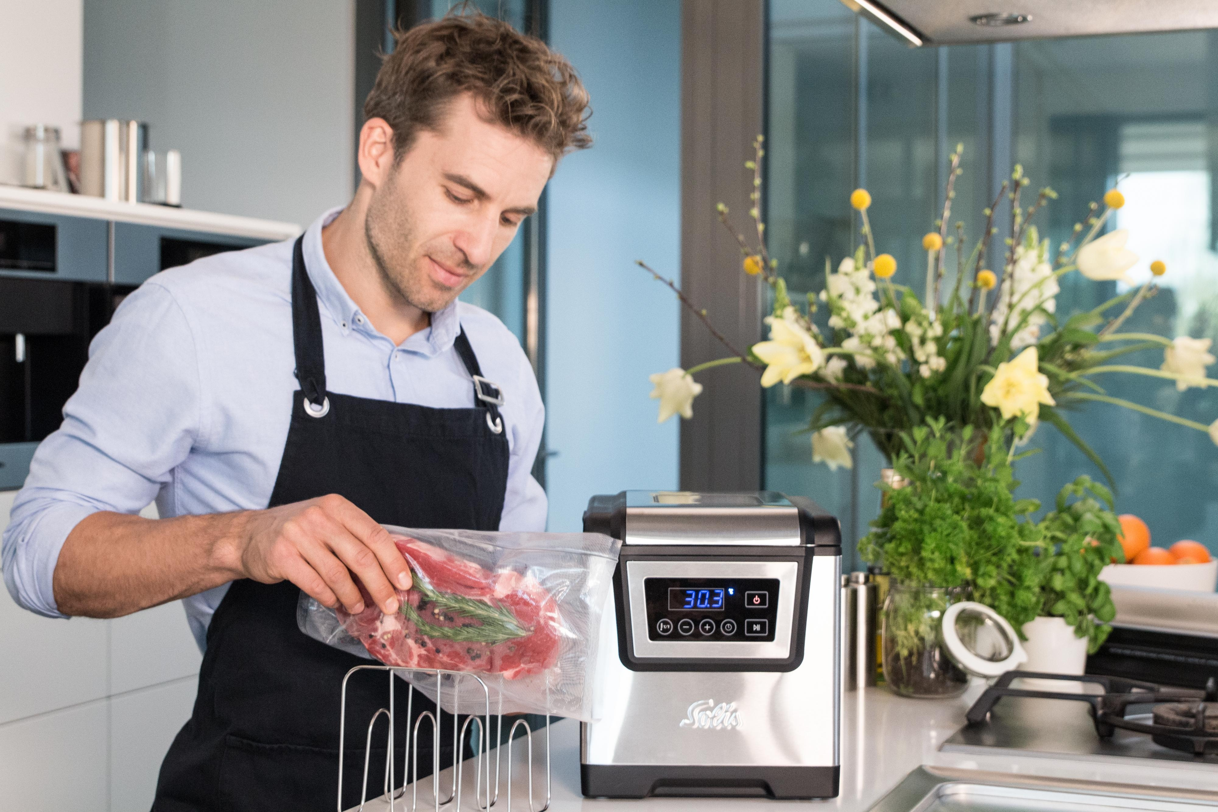Mann in Schürze legt Fleisch in einen Vakuumierer neben einem Sous-Vide-Gerät.