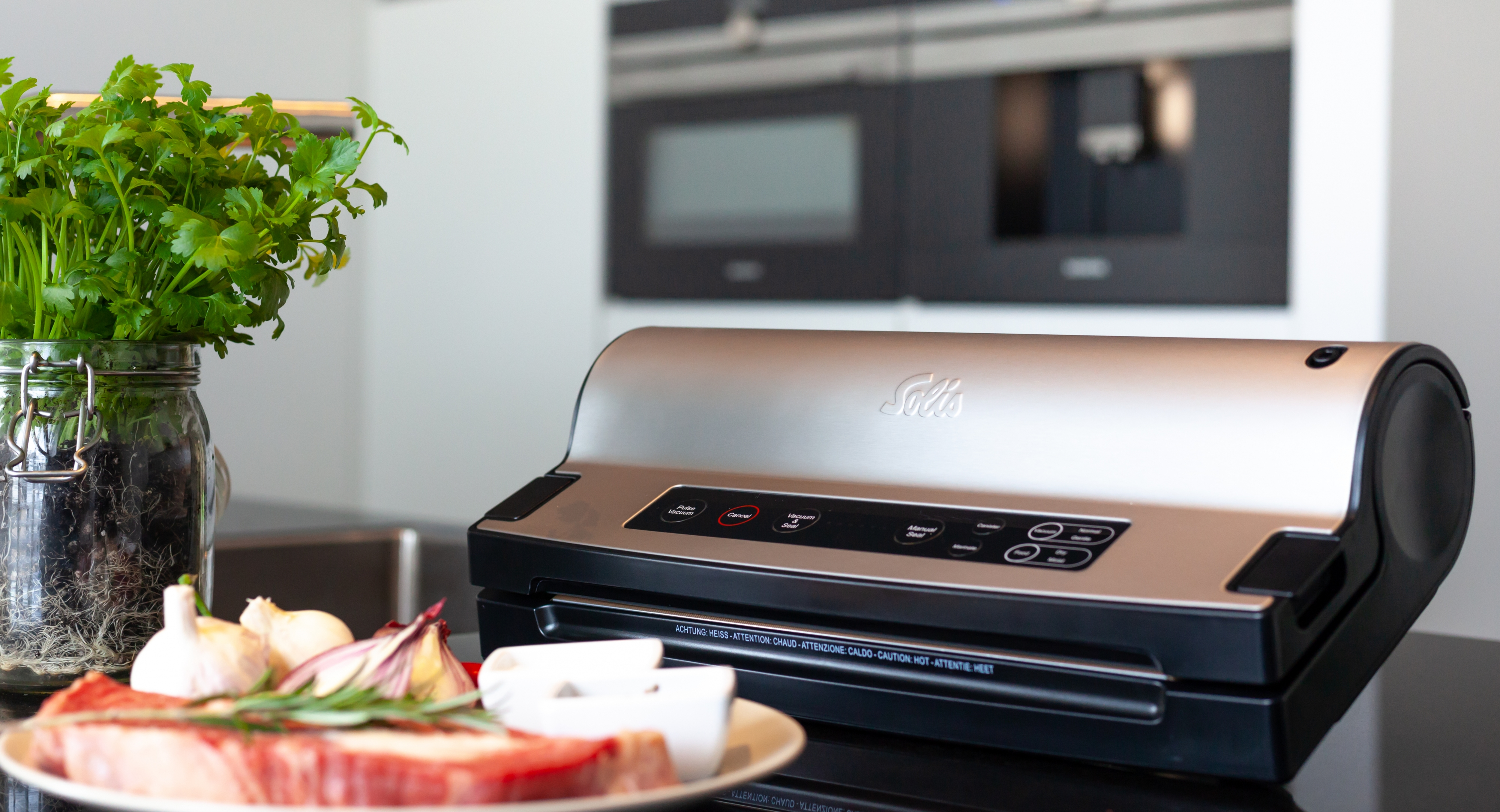 Zgrzewarka próżniowa obok produktów spożywczych na blacie kuchennym, z kuchnią w tle.