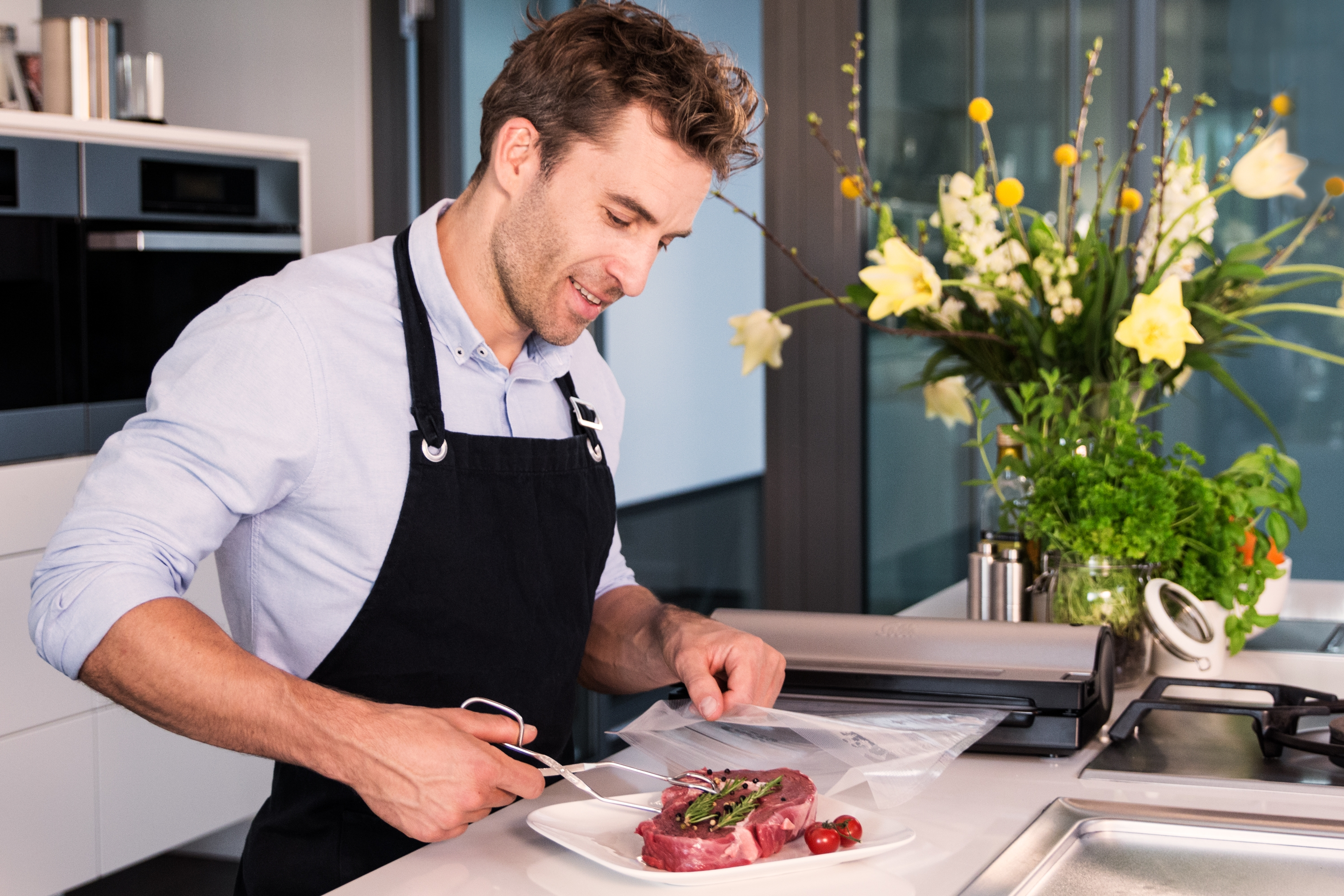 Mężczyzna w fartuchu kroi stek na talerzu, kwiaty w tle.