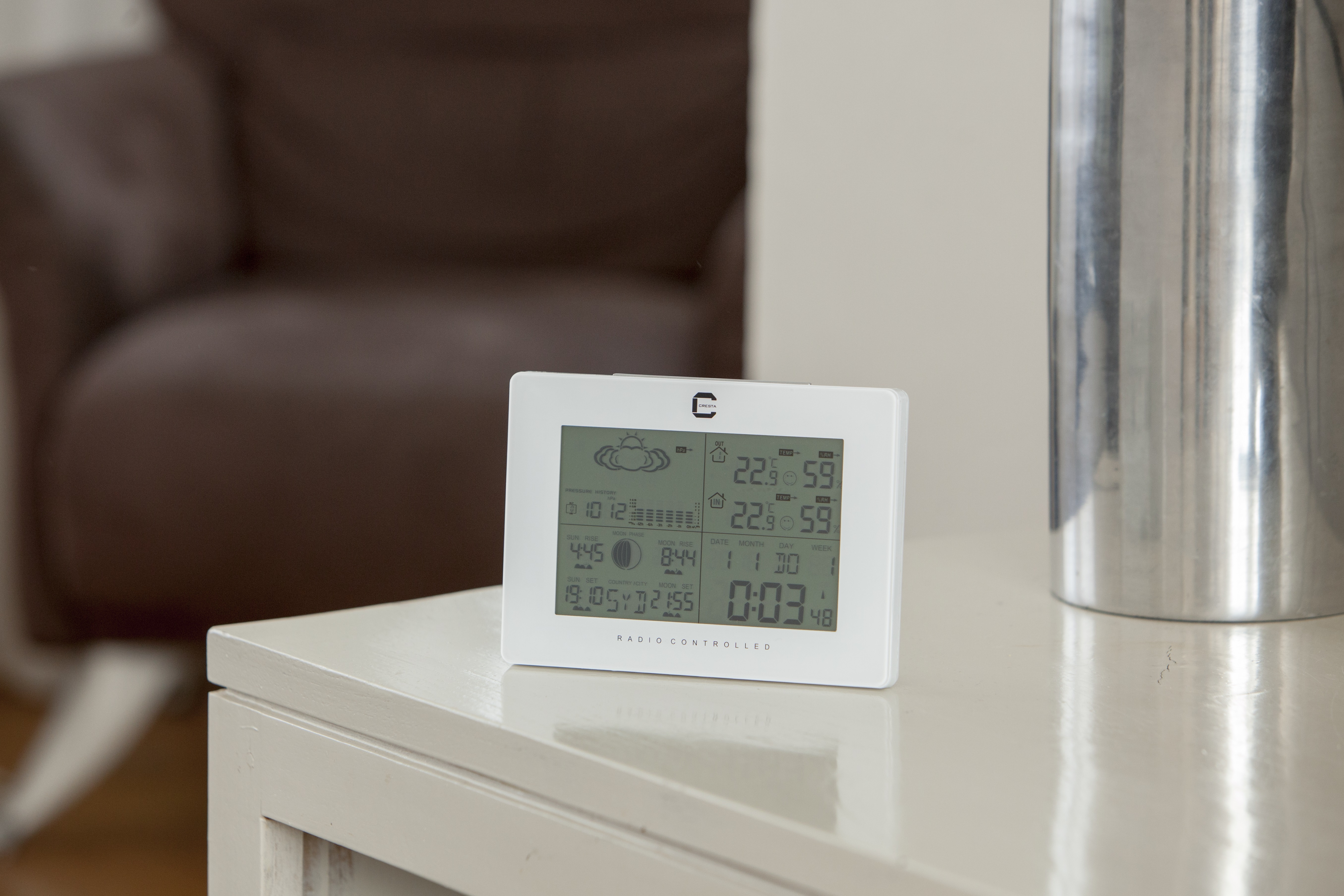 White weather station on a white table. It displays weather information. A sofa is blurred in the background.