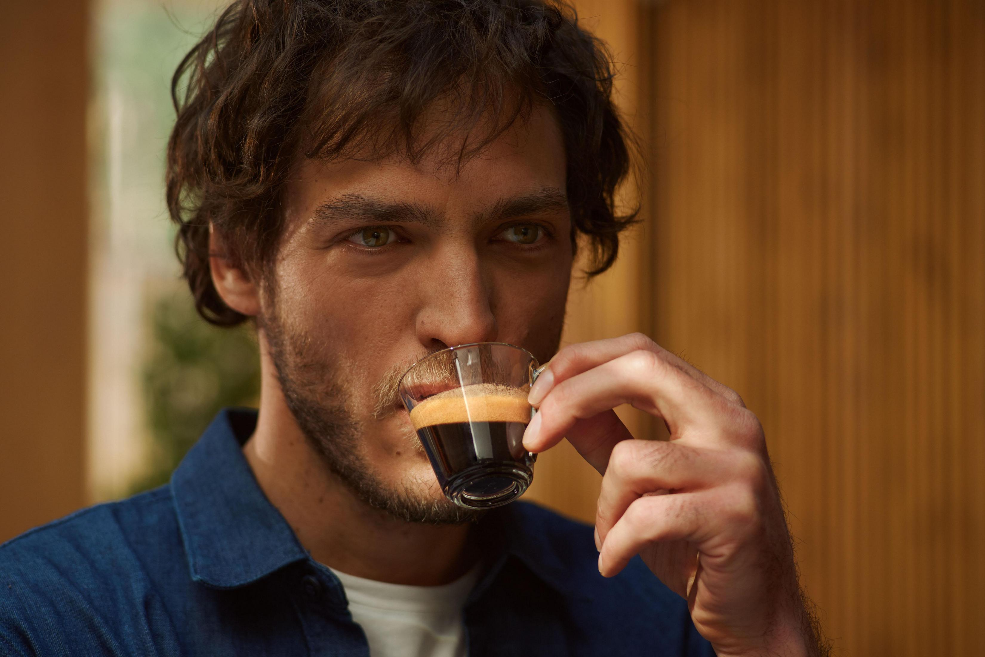 Ein Mann trinkt Espresso aus einem kleinen Glas. Er schaut in die Kamera. Der Hintergrund ist unscharf.