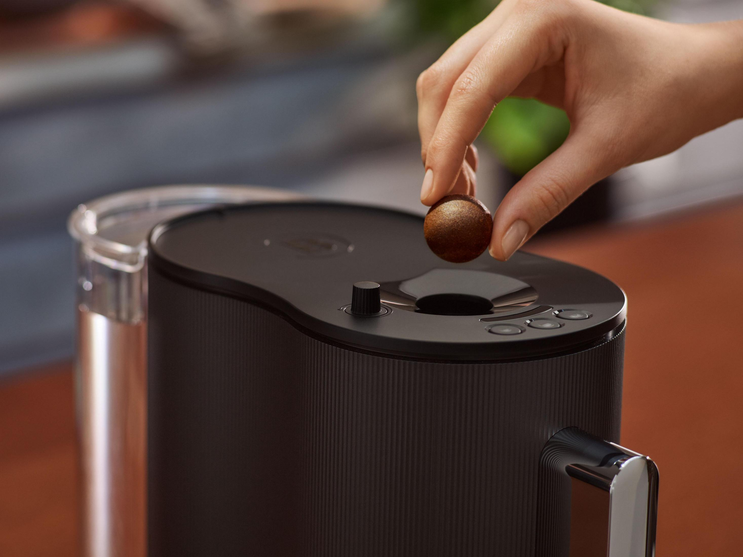 Eine Hand hält einen braunen Kaffee-Pod über einer schwarzen Kaffeemaschine.