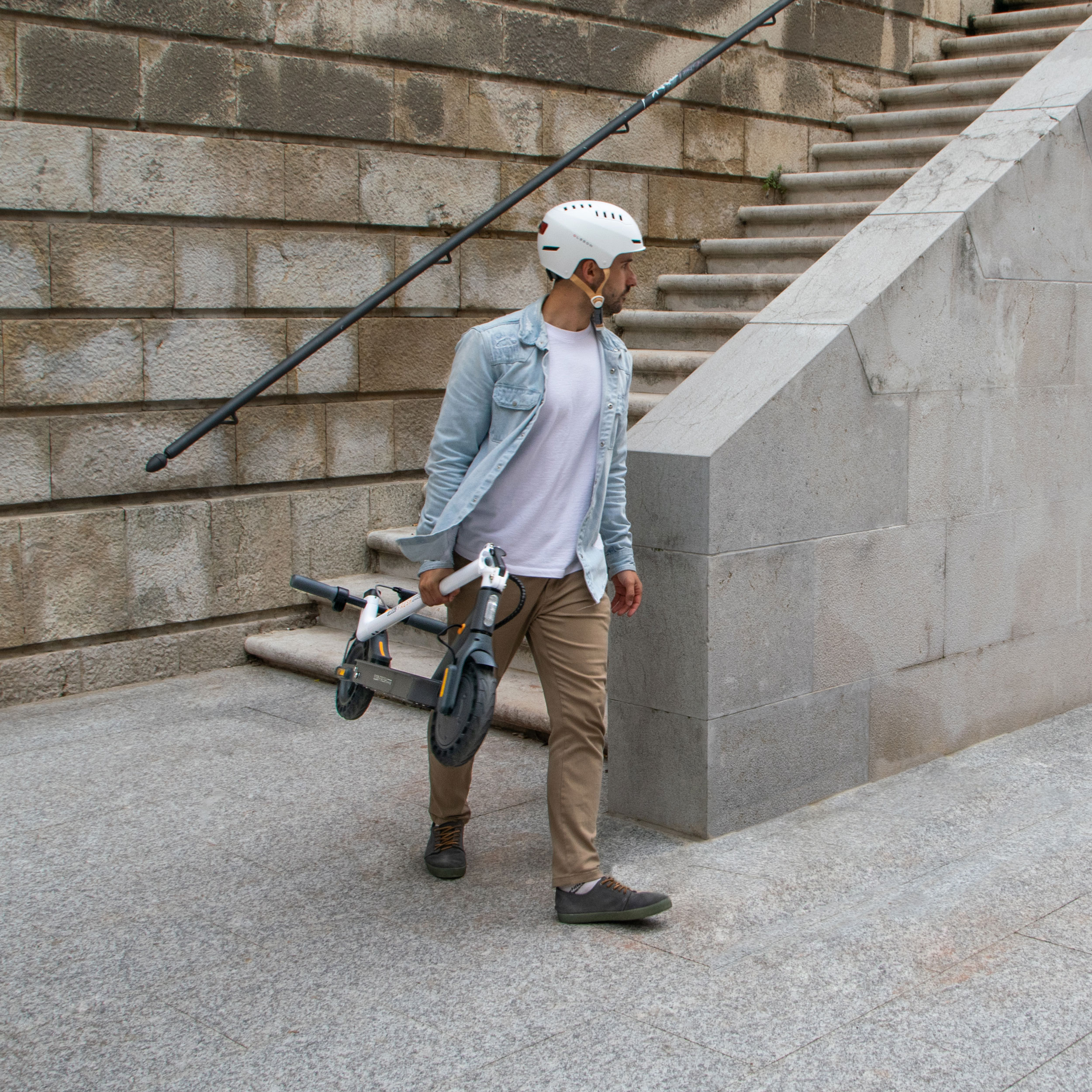 Mann trägt zusammengeklappten Roller. Er trägt Helm, Hemd und Hose. Treppe und Wand im Hintergrund.