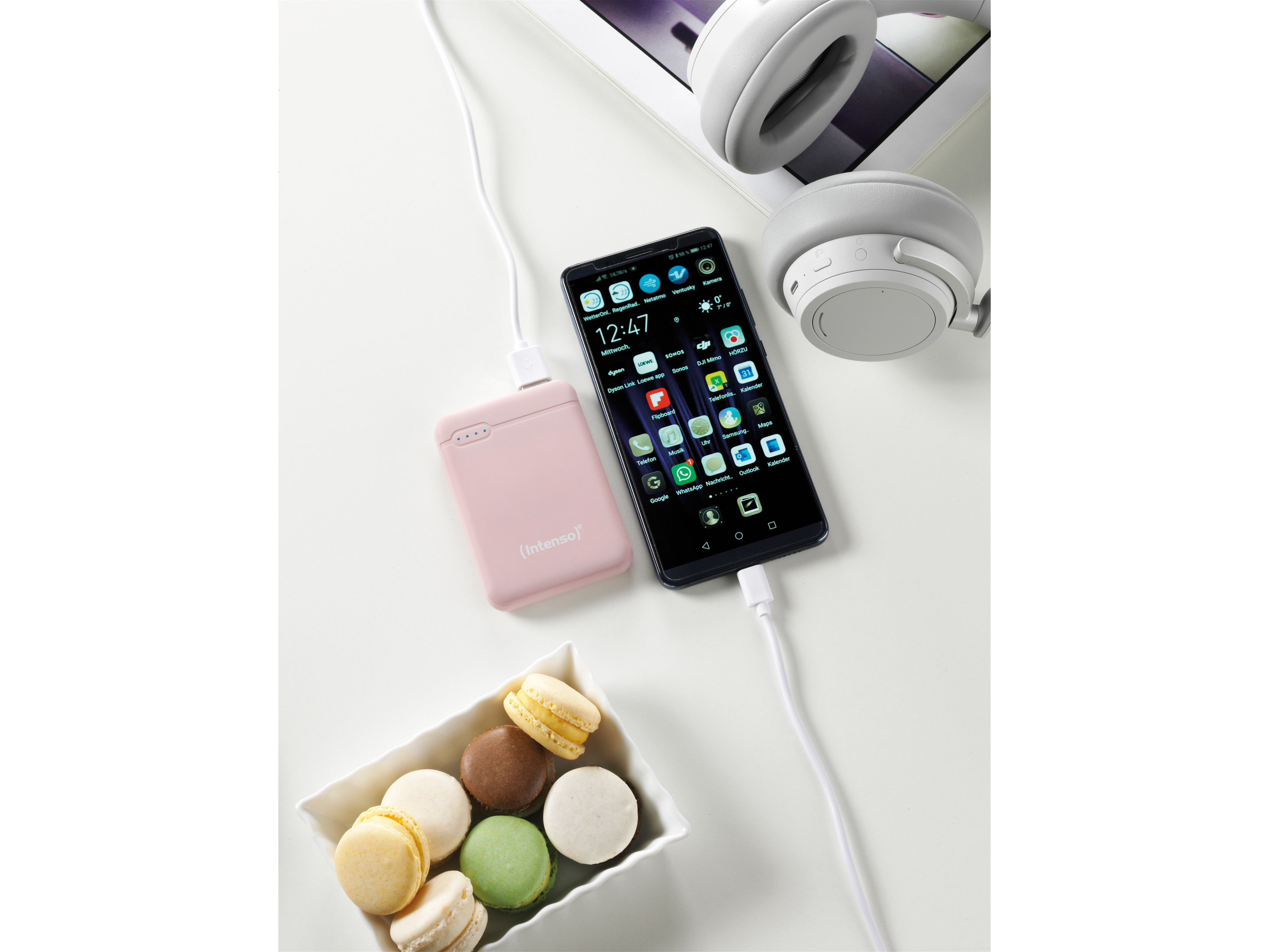 Ein Telefon, das von einer pinken Powerbank geladen wird, mit Makronen und Kopfhörern auf einem Tisch.