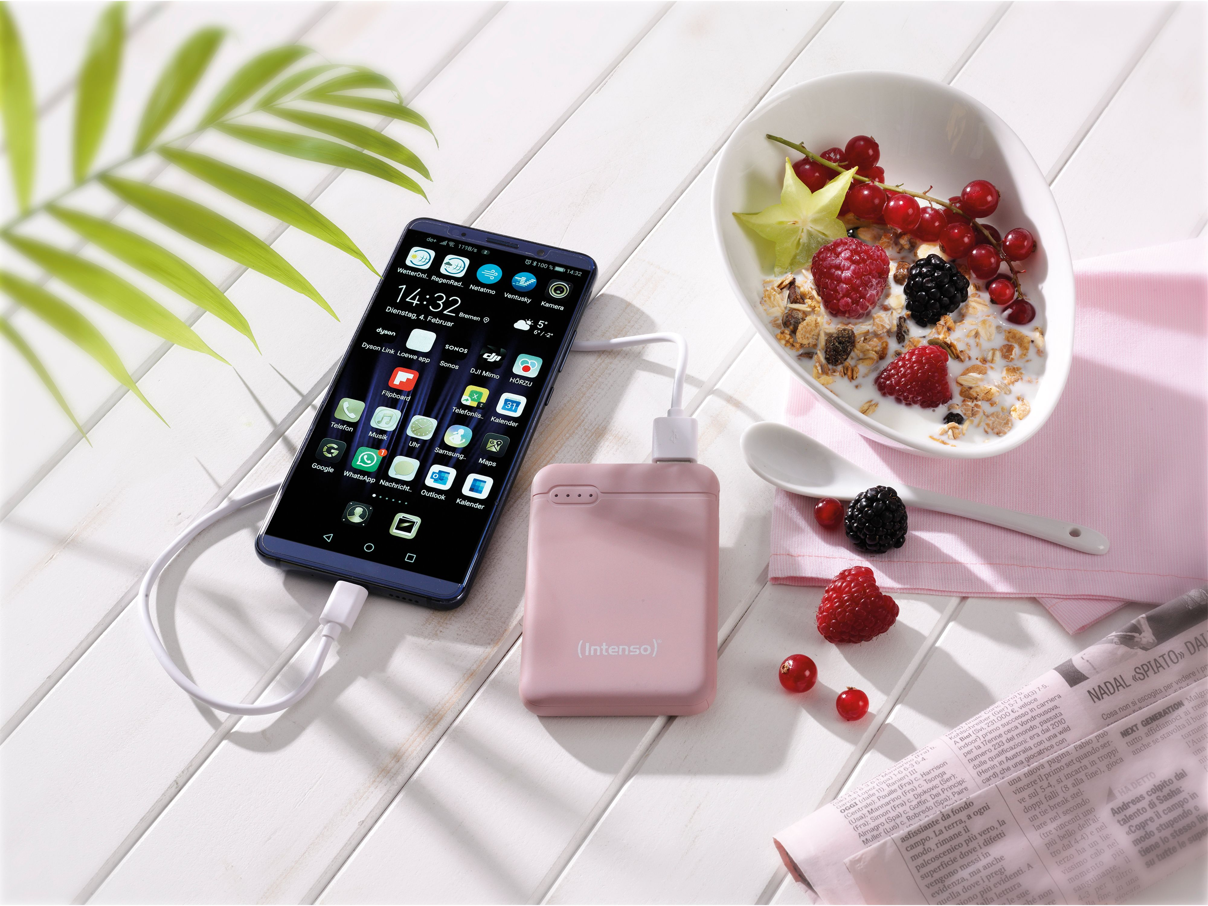 Ein Telefon, das mit einer rosa Powerbank aufgeladen wird, neben einer Schüssel mit Beeren und einer Zeitung.