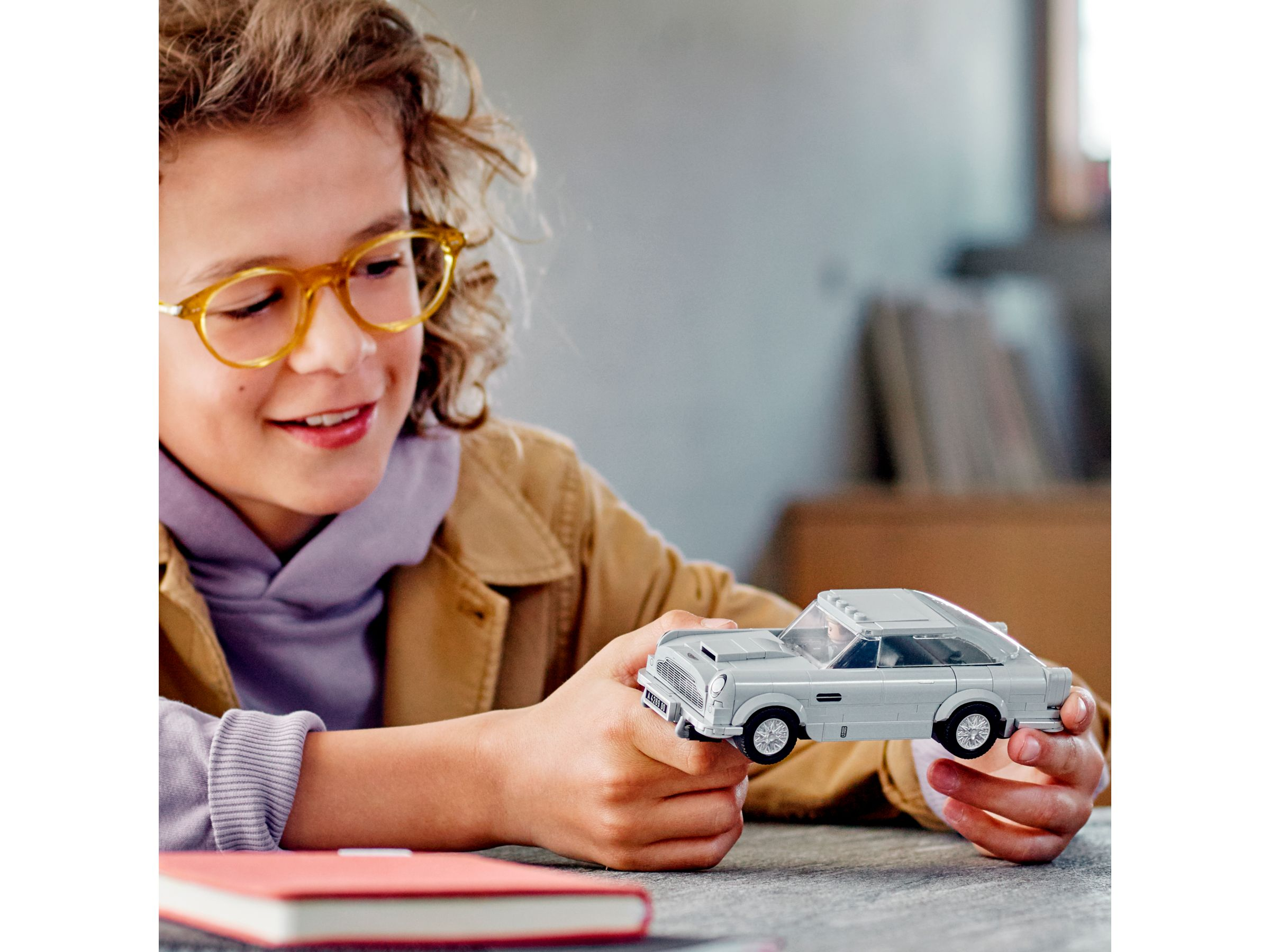 Ein Junge hält ein silbernes Lego-Auto. Er trägt eine Brille, eine braune Jacke und ein lila Hemd.