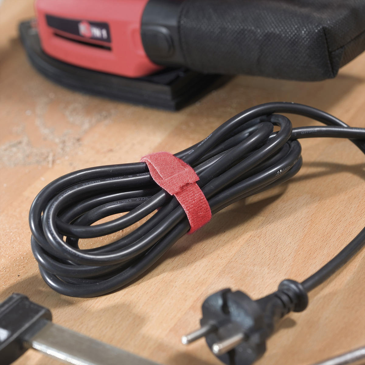 Cavo elettrico nero avvolto con una chiusura a strappo rossa. Un elettroutensile è sullo sfondo.