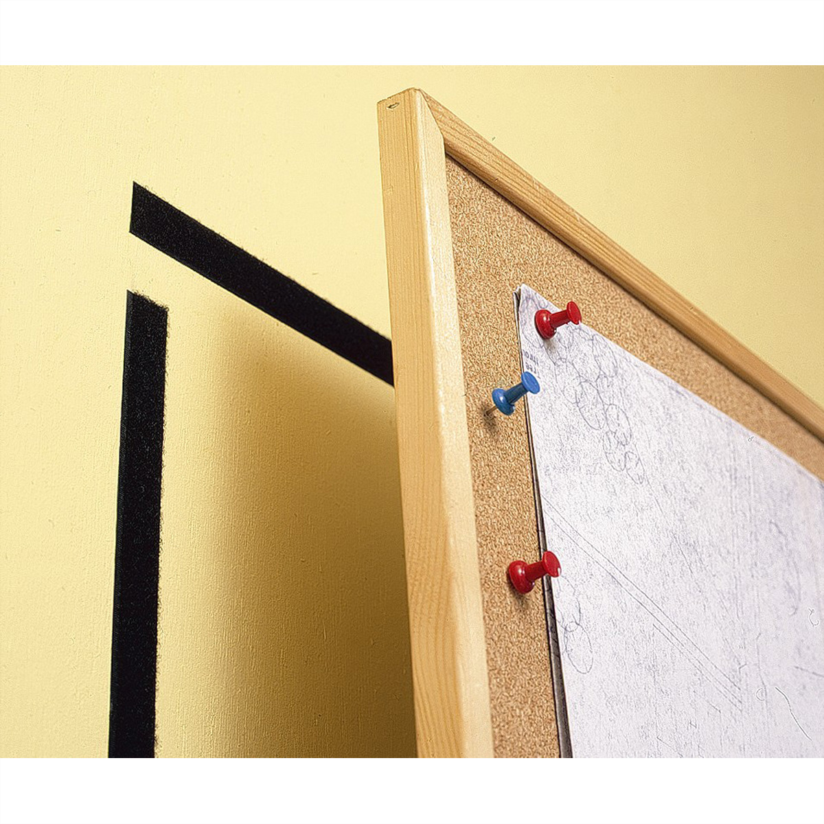 Bacheca in sughero con disegno appuntato da puntine rosse e blu, su muro giallo con nastro nero.