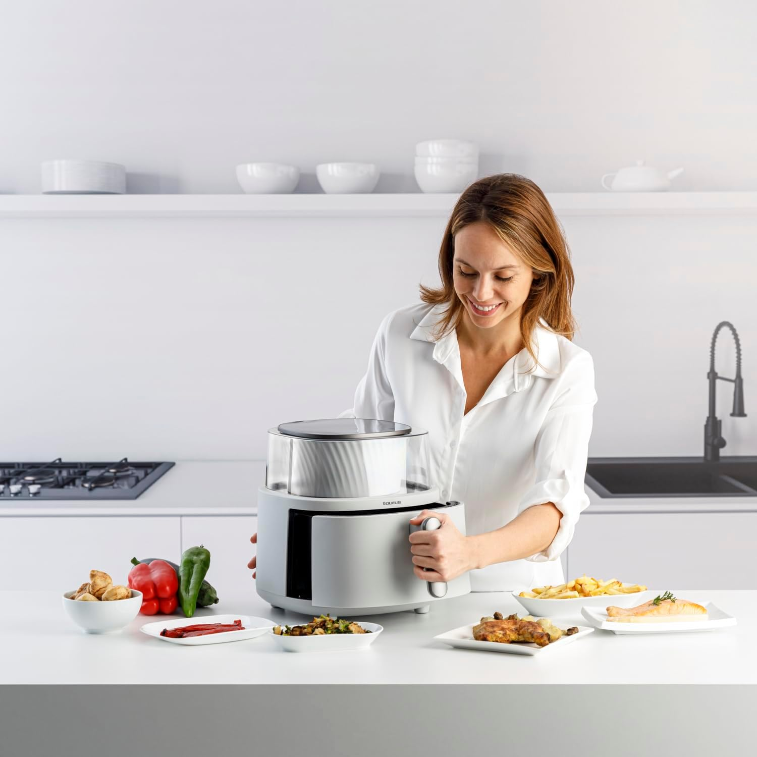 Mujer sonriendo usando un electrodoméstico plateado con platos de comida en la encimera. Cocina blanca con objetos.