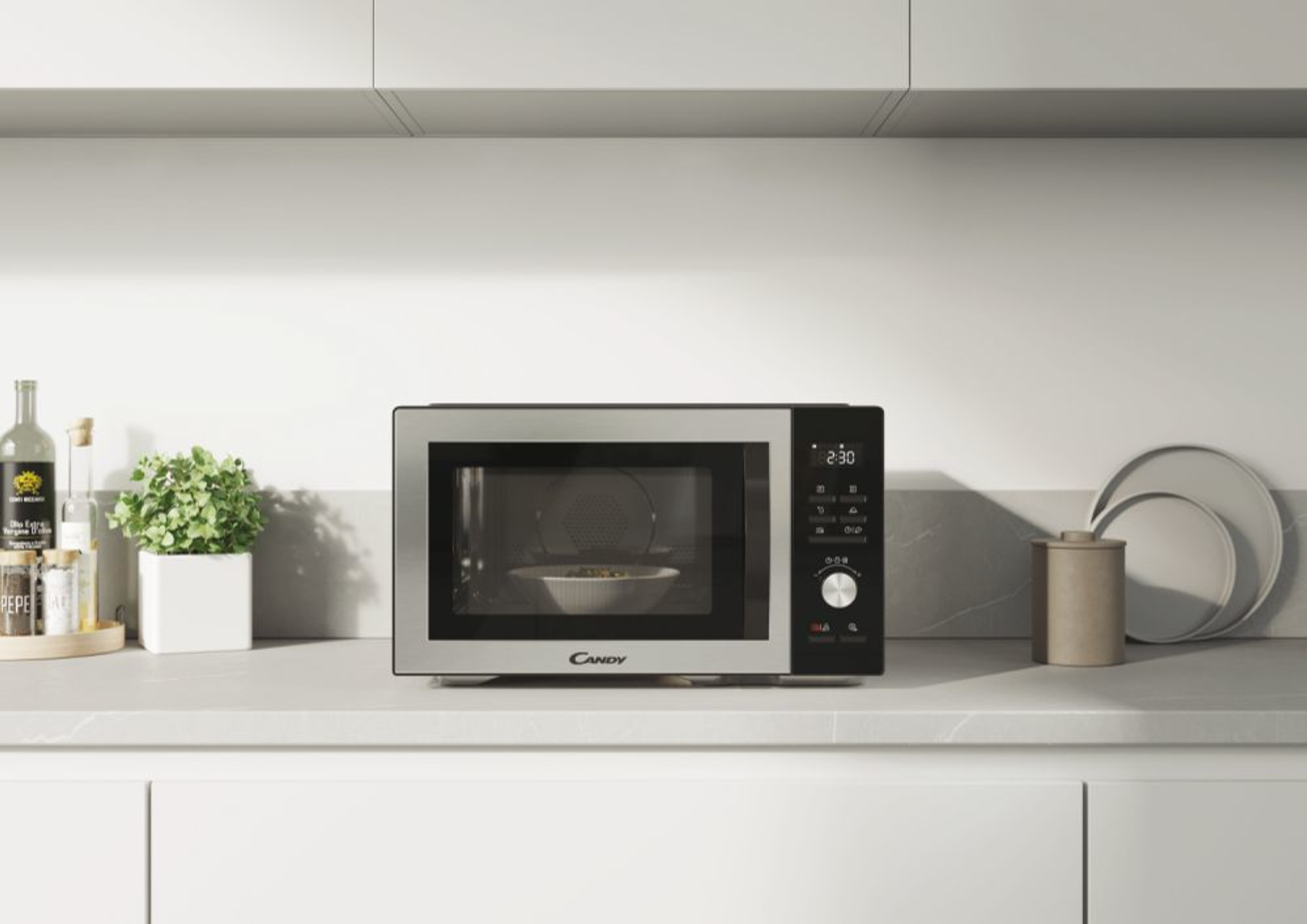 Kuchenka mikrofalowa na białym blacie z rośliną, butelkami i talerzem w tle.
