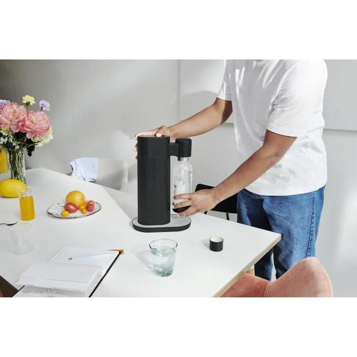 Eine Person fügt einer Soda-Maschine eine Flasche hinzu. Auf dem Tisch befinden sich Blumen, Obst und Wasser.