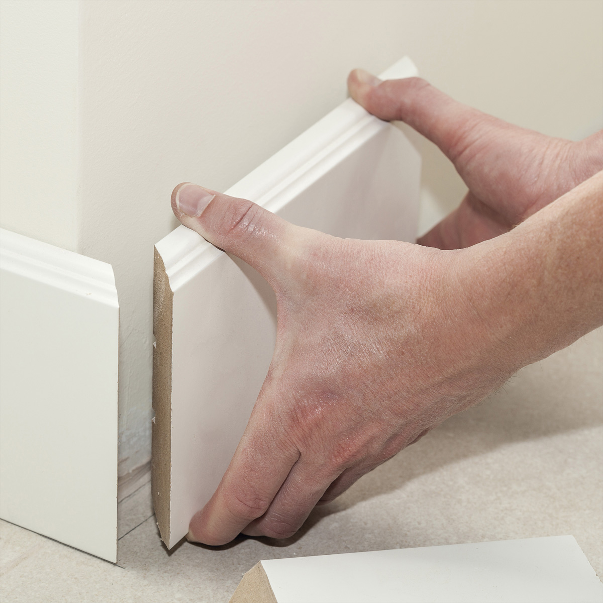 Person installiert weiße Fußleiste an einer Wand. Hände halten die Leiste, weiße Wand und Boden.