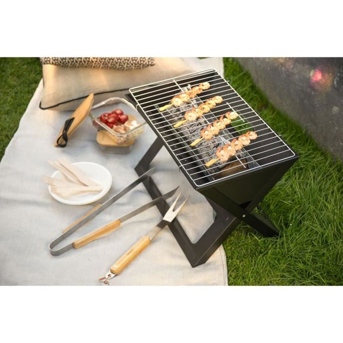 Kleiner tragbarer Grill mit Spießen mit Essen, das auf einer grasbewachsenen Oberfläche mit Utensilien in der Nähe gegart wird.