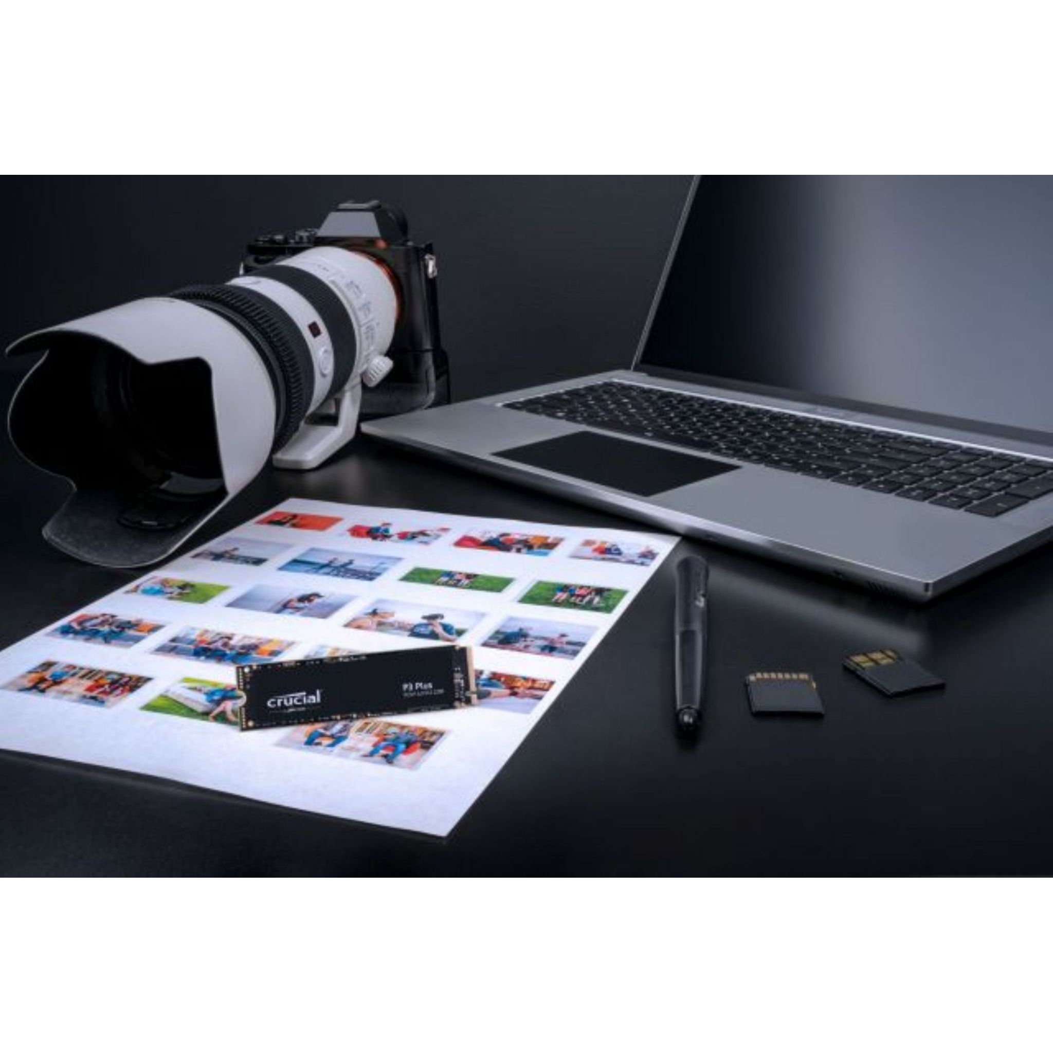 Laptop, aparat fotograficzny, dysk SSD Crucial i długopis na ciemnej powierzchni, z wydrukami zdjęć.