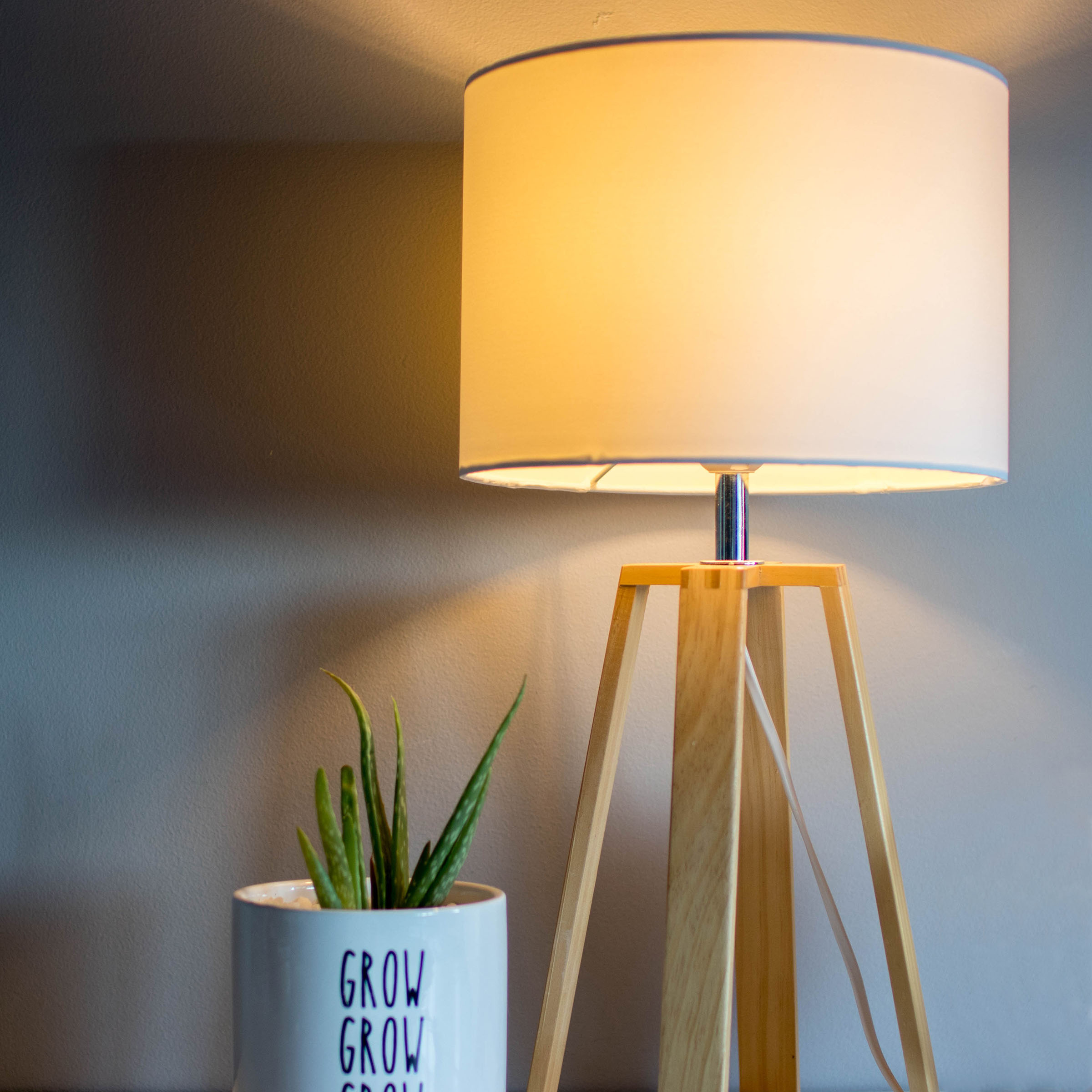 Stehlampe mit weißem Schirm und Holzbeinen. Eine kleine Topfpflanze befindet sich in der Nähe.