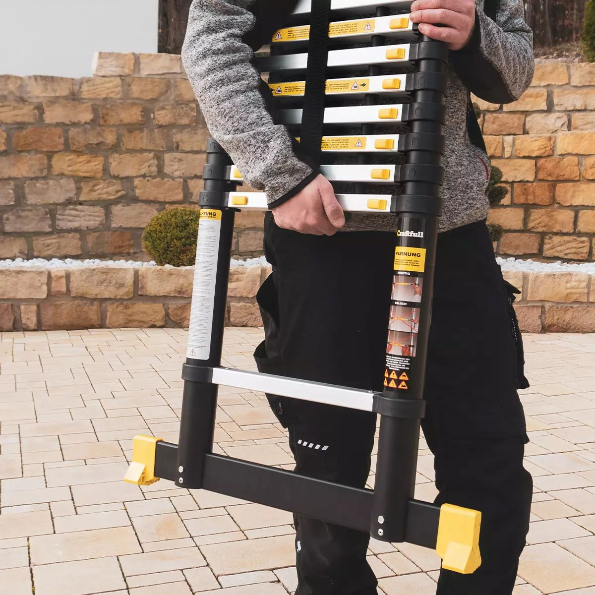 Ein Mann hält eine schwarz-gelbe Teleskopleiter. Er trägt einen grauen Pullover und schwarze Hose.