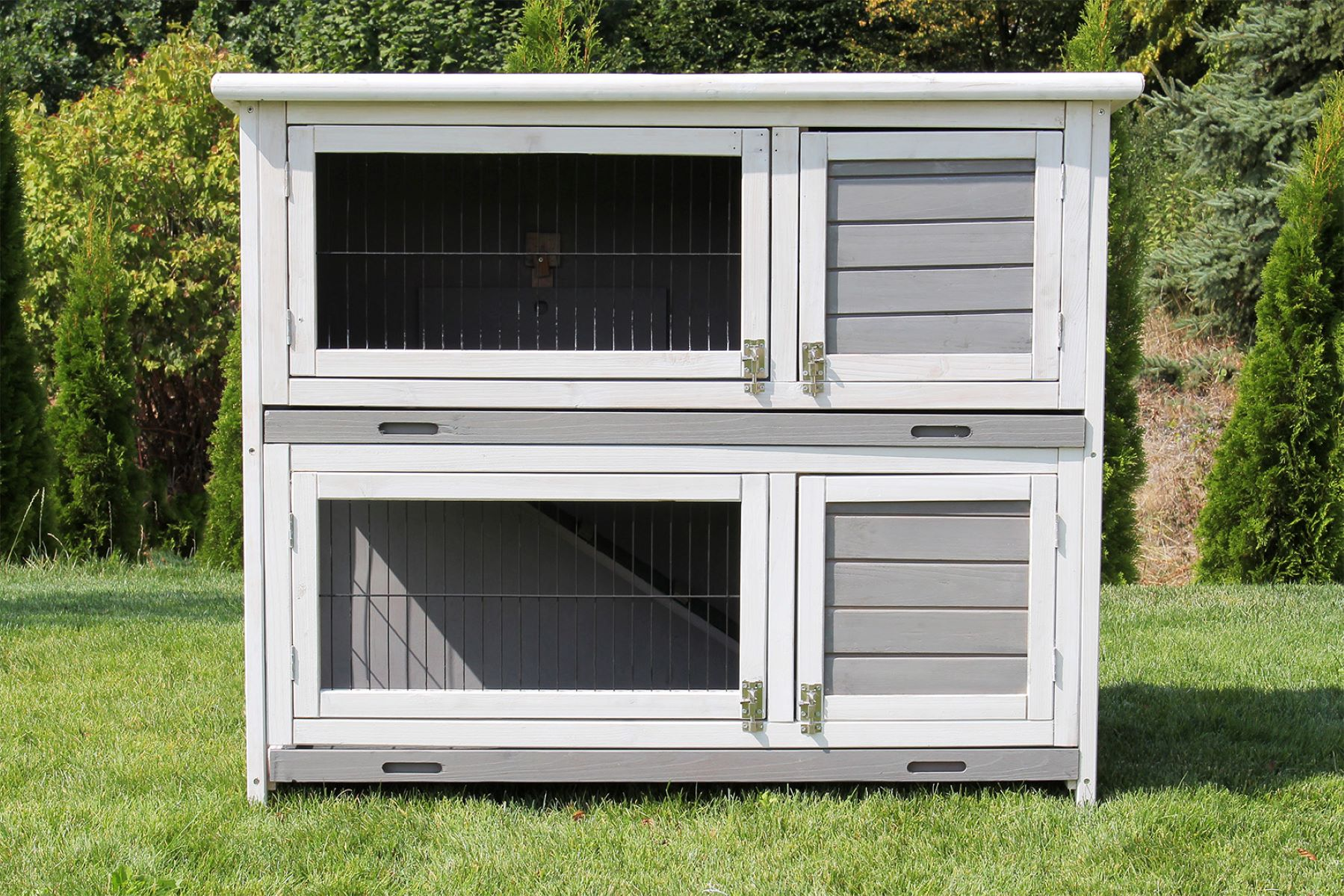 Ein zweistöckiger Kaninchenstall aus Holz mit Drahtgeflecht-Türen, auf Gras stehend, mit Bäumen im Hintergrund.