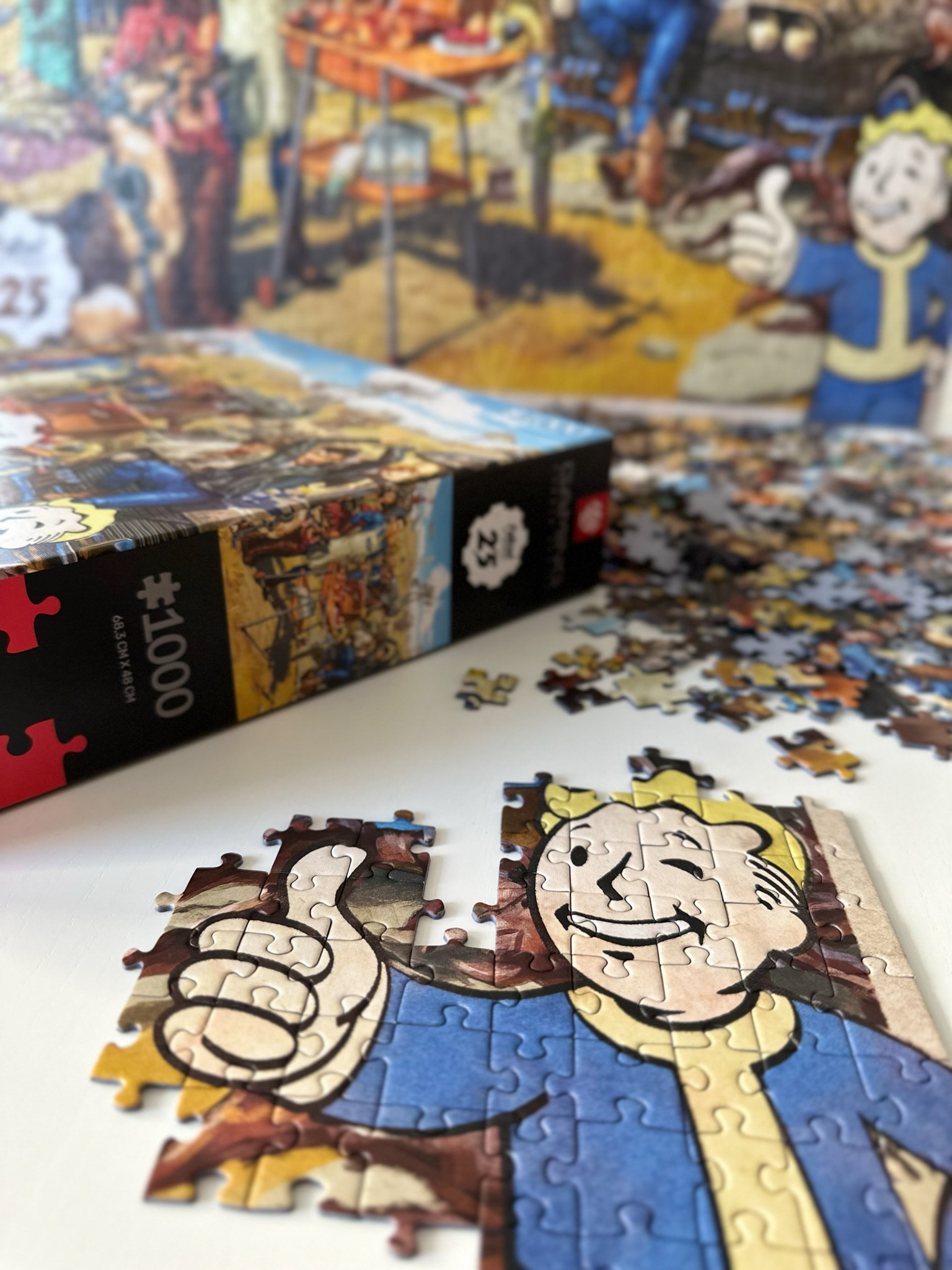 Puzzle z postacią Vault Boy, kciuk w górę. Pudełko i rozrzucone elementy. Tło z obrazem targu.