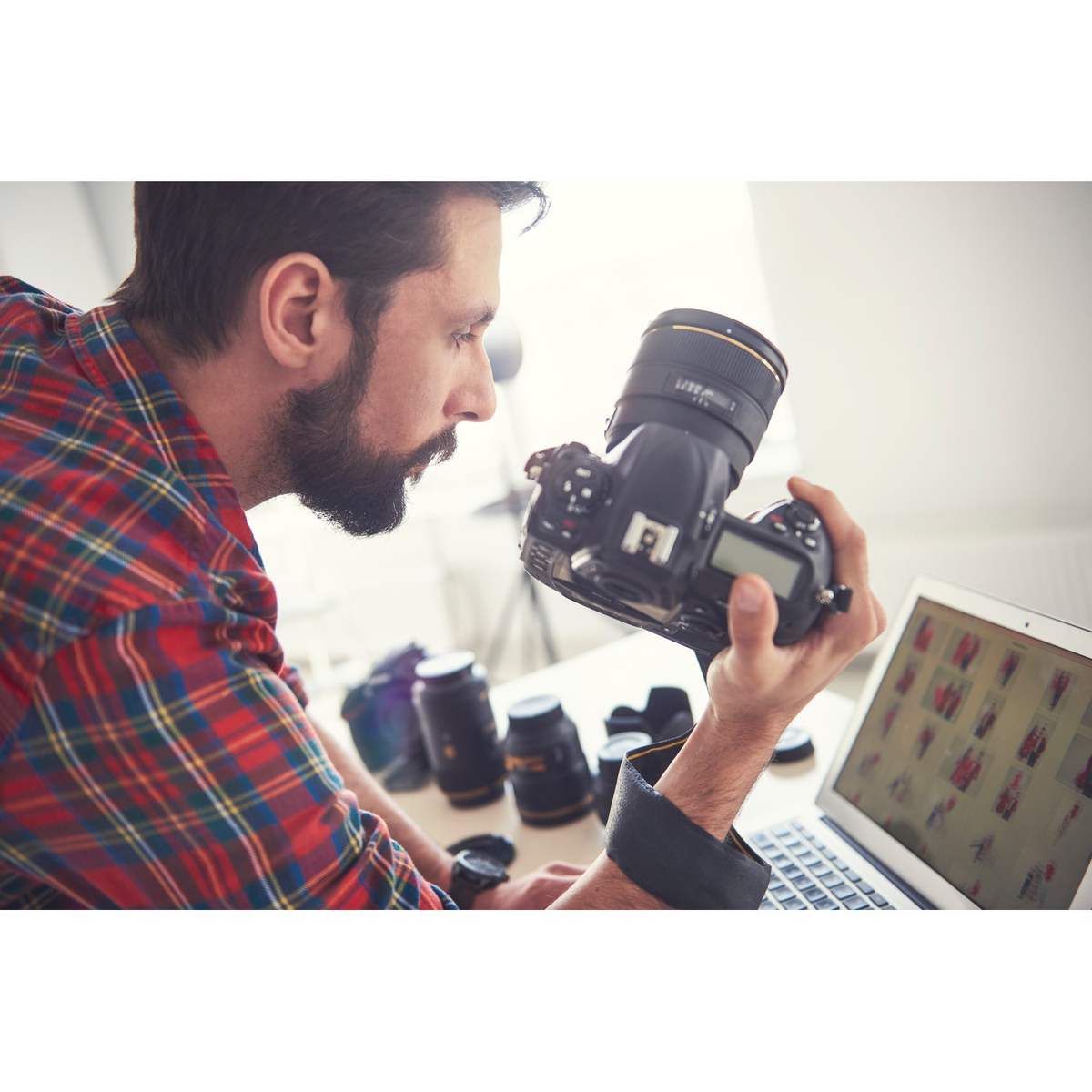 Hombre con barba examina una cámara en un escritorio con lentes y un portátil mostrando fotos.