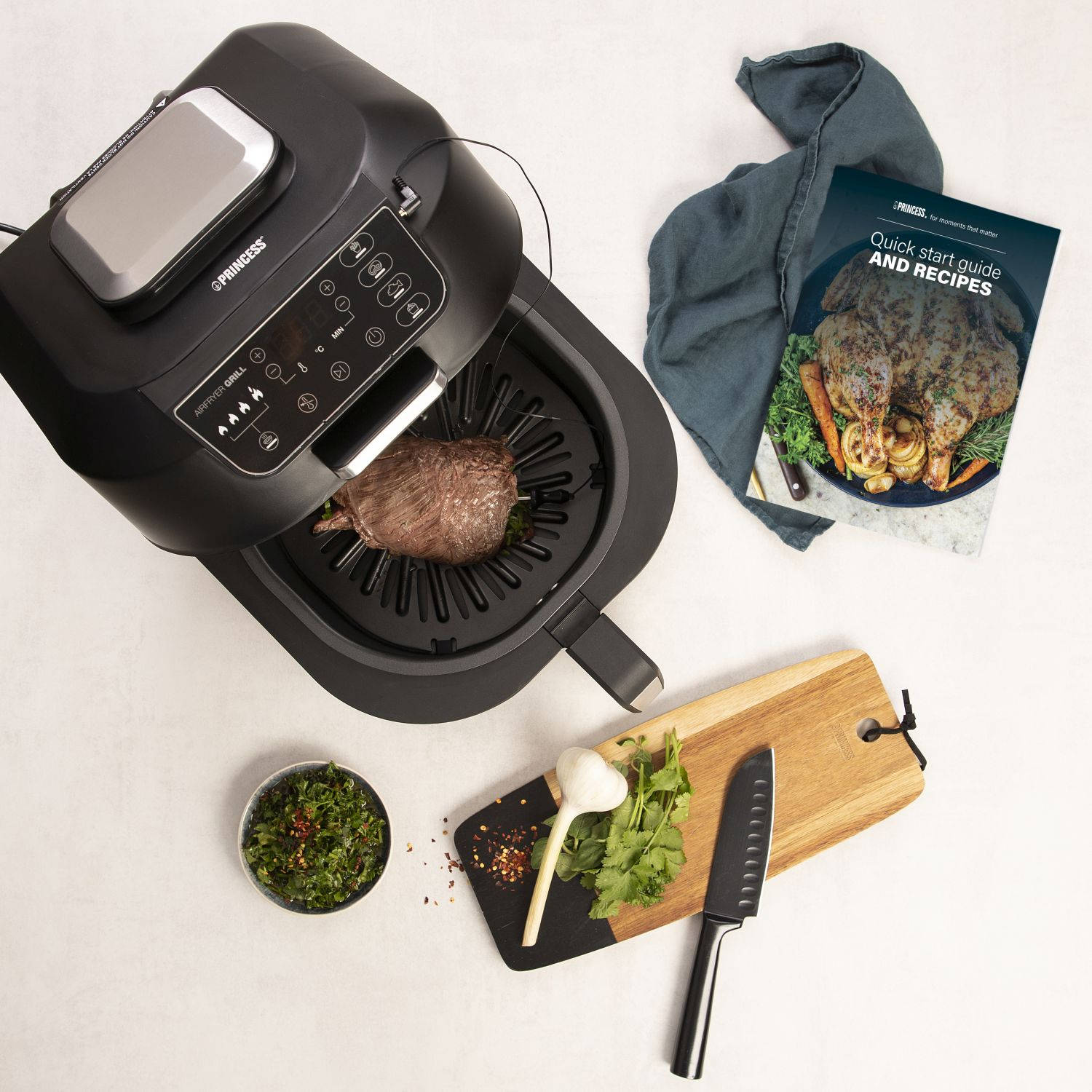 Frytownica powietrzna z włożonym stekiem, deska do krojenia, nóż, zioła i książka z przepisami.