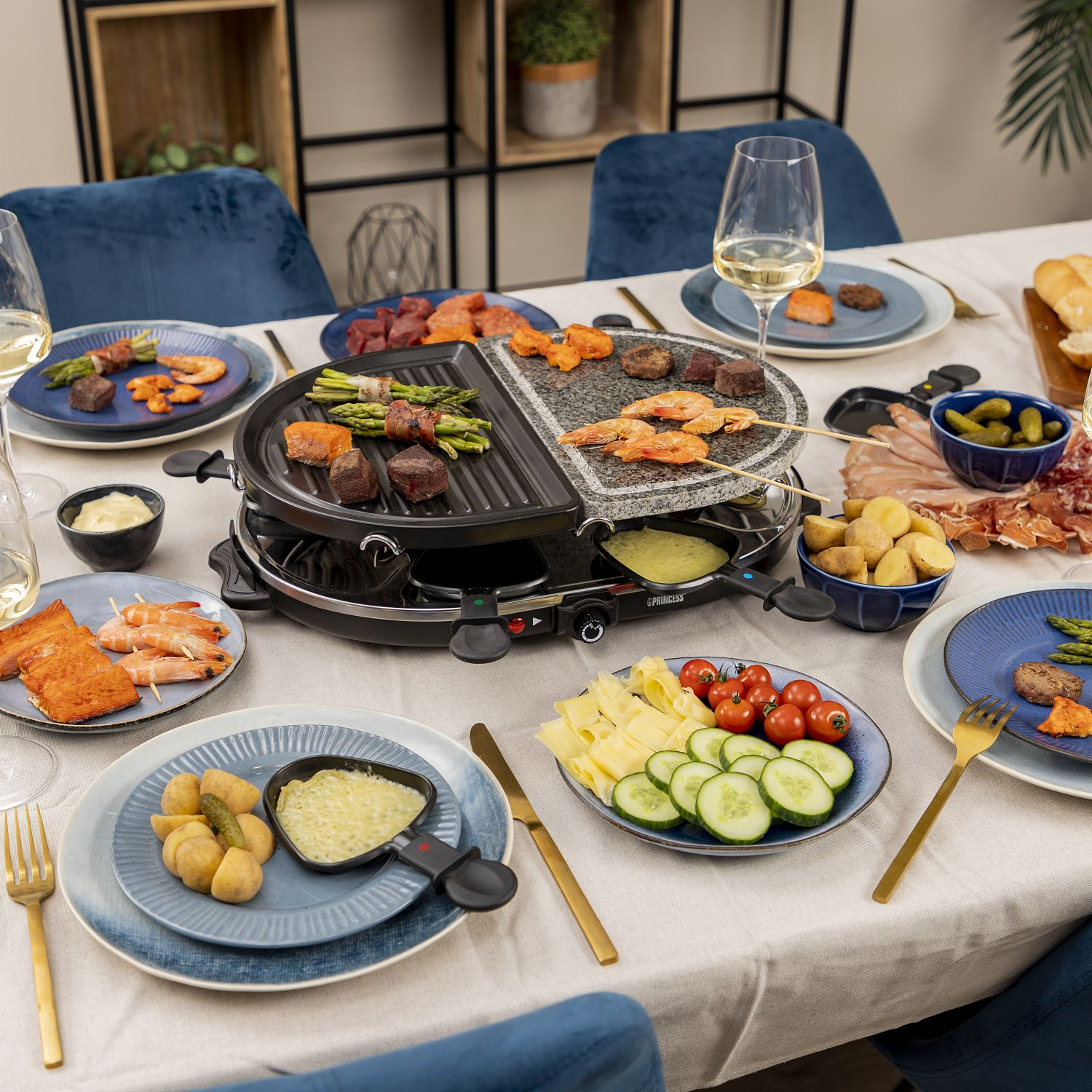 Nakrycie stołu z grillem do raclette, różnymi produktami spożywczymi, talerzami i szklankami.