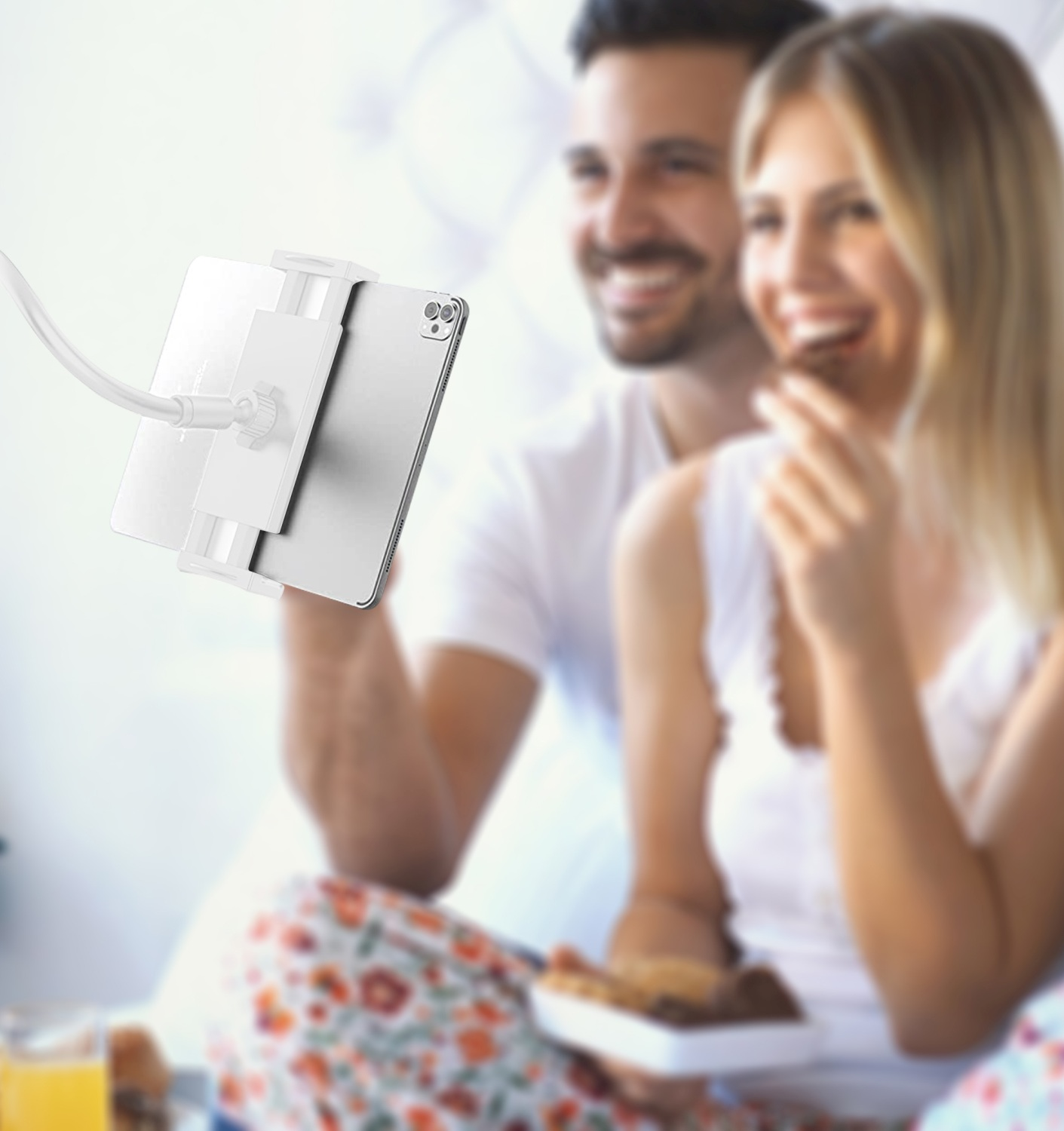 Paar im Bett mit Tablet-Ständer. Tablet, Tisch und Personen sichtbar. Weiße Bettwäsche.