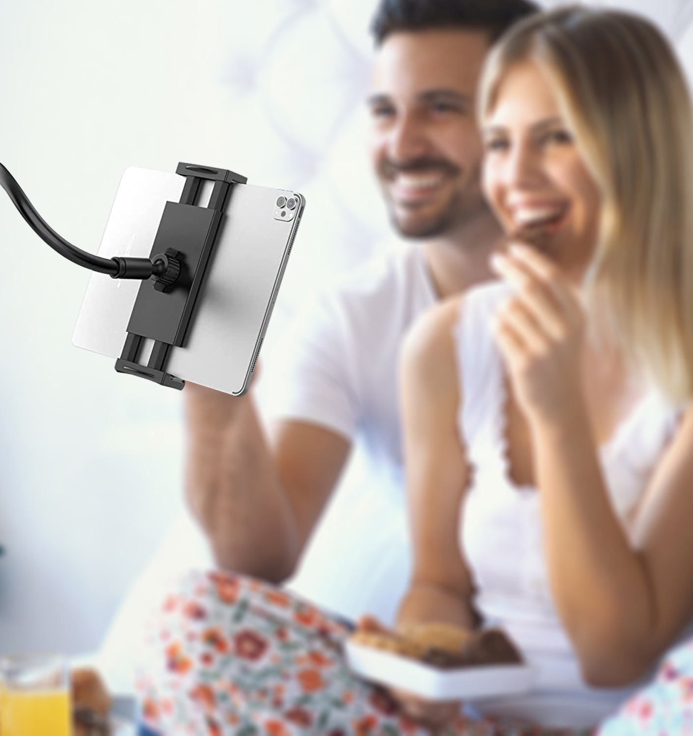 Ein lächelndes Paar, das auf ein Tablet schaut, das von einer flexiblen Halterung gehalten wird. Sie essen Snacks.