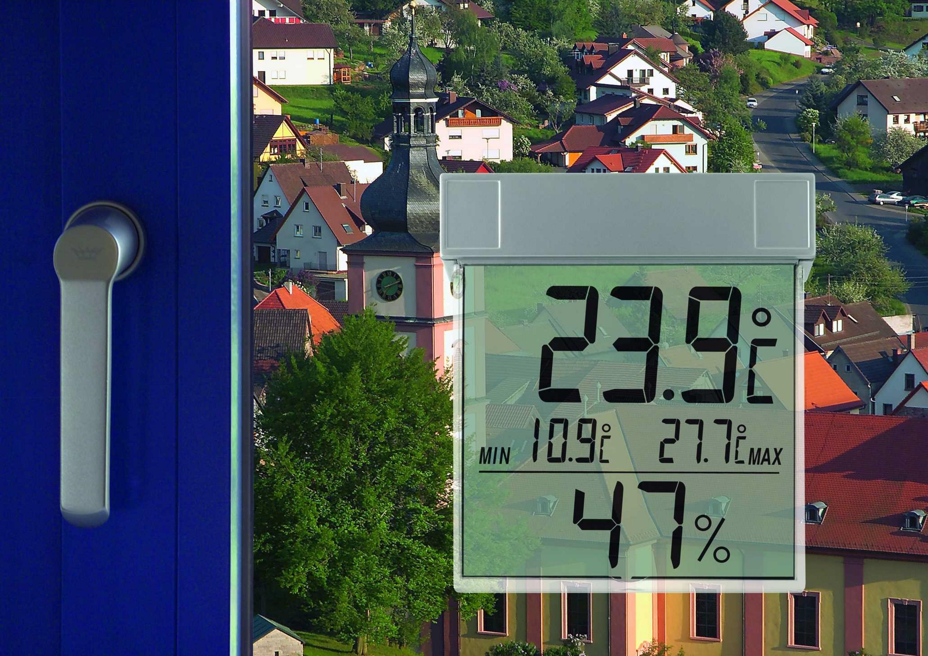 Ein digitales Thermometer zeigt 23,9°C, mit min/max Temperaturen und 47% Luftfeuchtigkeit. Im Hintergrund eine Kirche und Häuser.