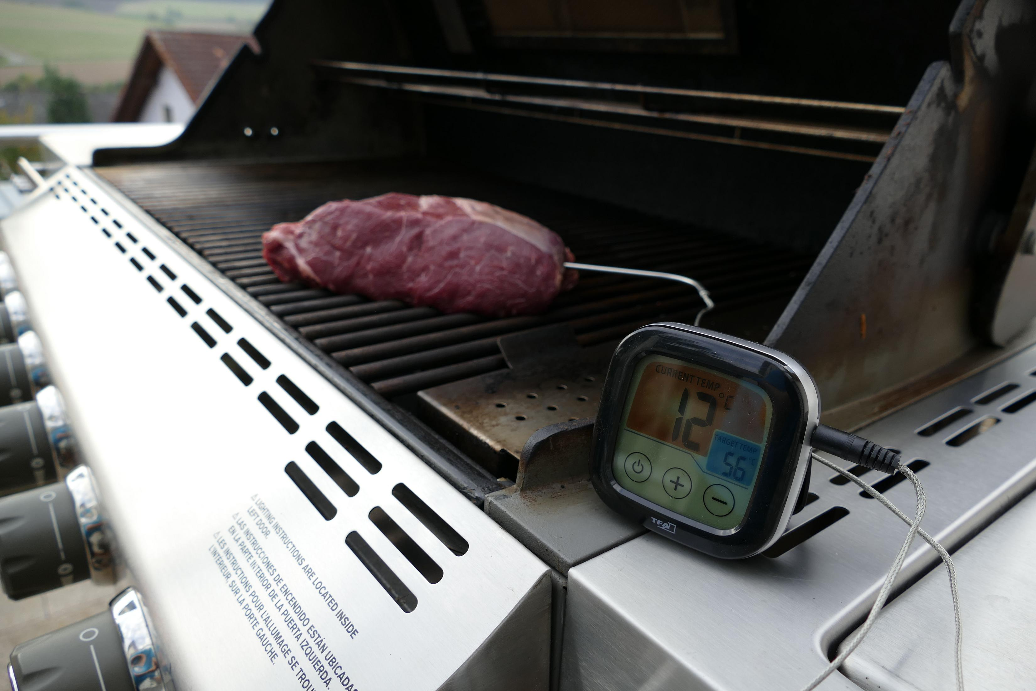 Ein Grill mit Fleisch und Thermometer. Im Hintergrund ein Haus und Bäume.