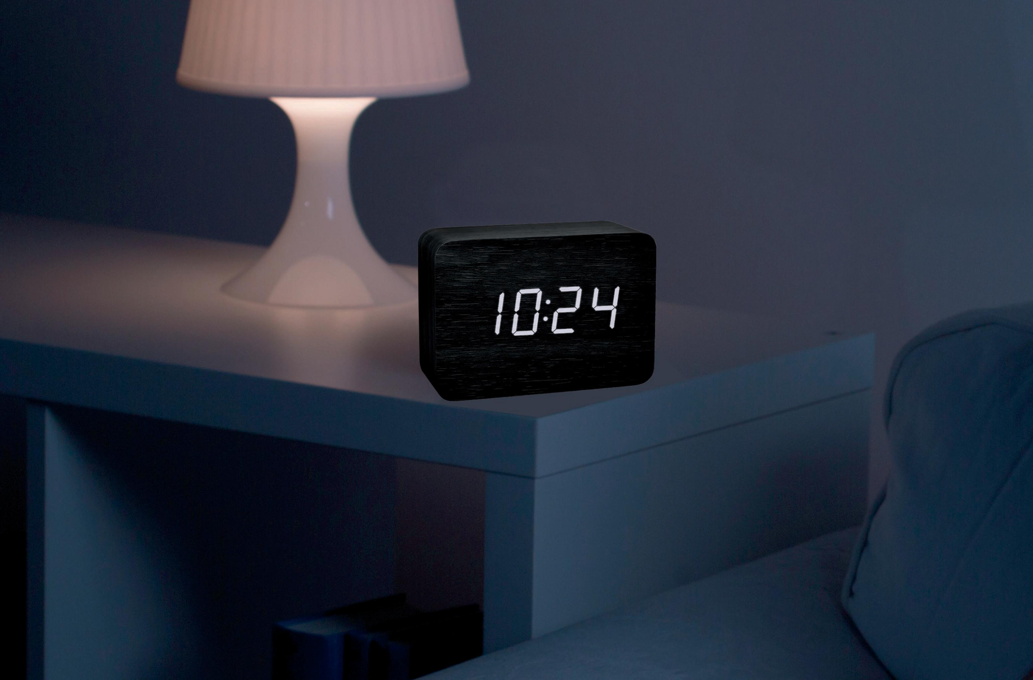 Schwarzer Wecker aus Holz, der 10:24 anzeigt, auf einem weißen Tisch. Im Hintergrund eine Lampe.