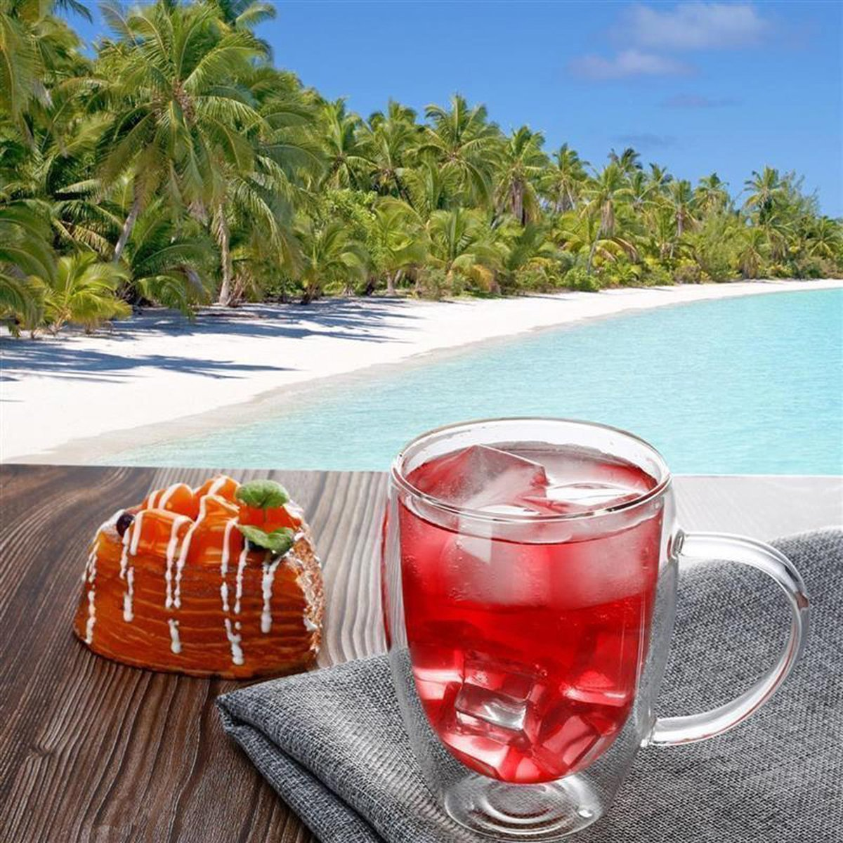 Strandszene mit Palmen, weißem Sand und türkisfarbenem Wasser. Ein Glas rotes Getränk mit Eis und Gebäck.