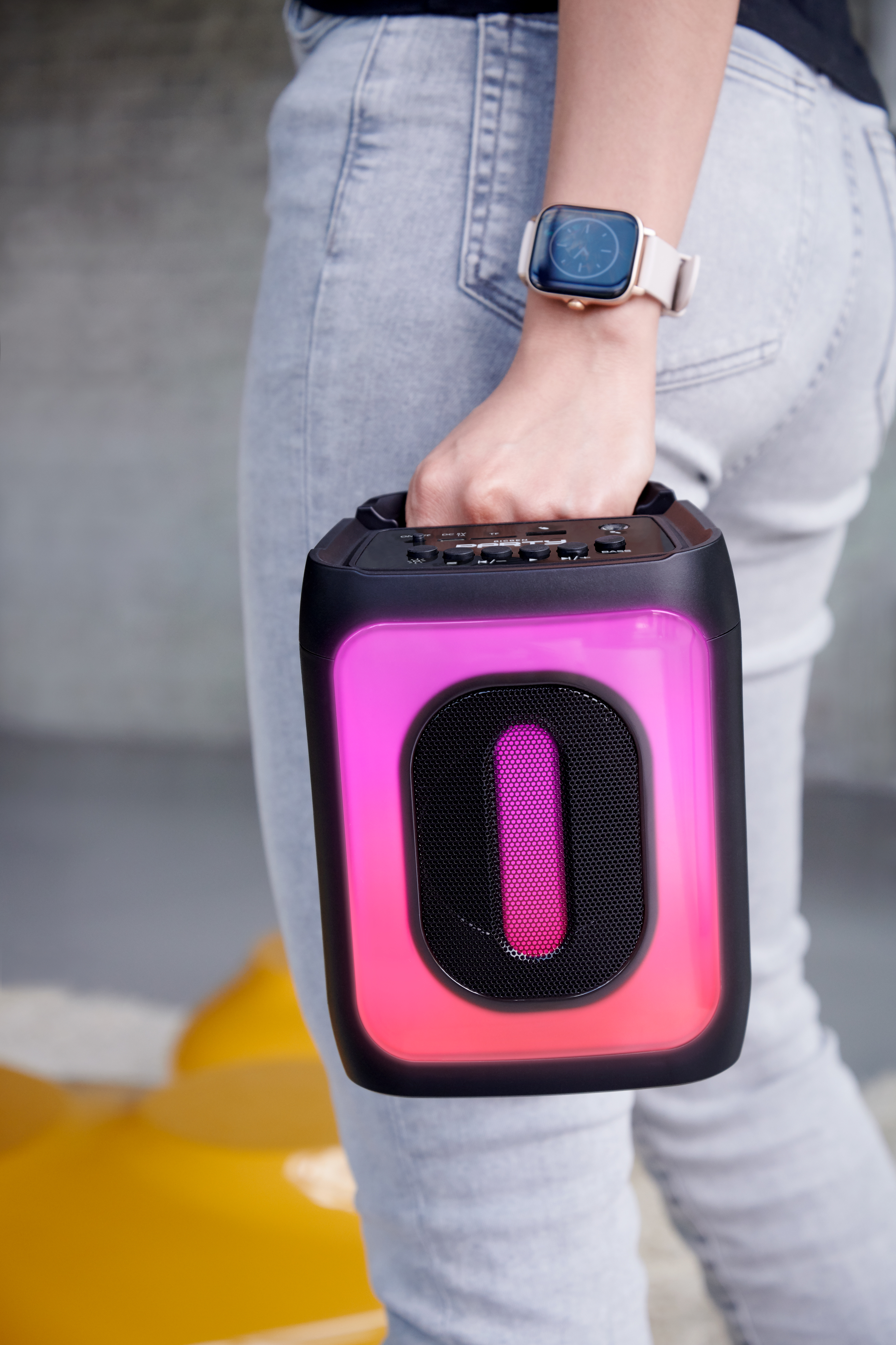 Osoba trzyma głośnik przenośny. Głośnik ma różowe światła i czarny uchwyt. Osoba ma na sobie dżinsy.