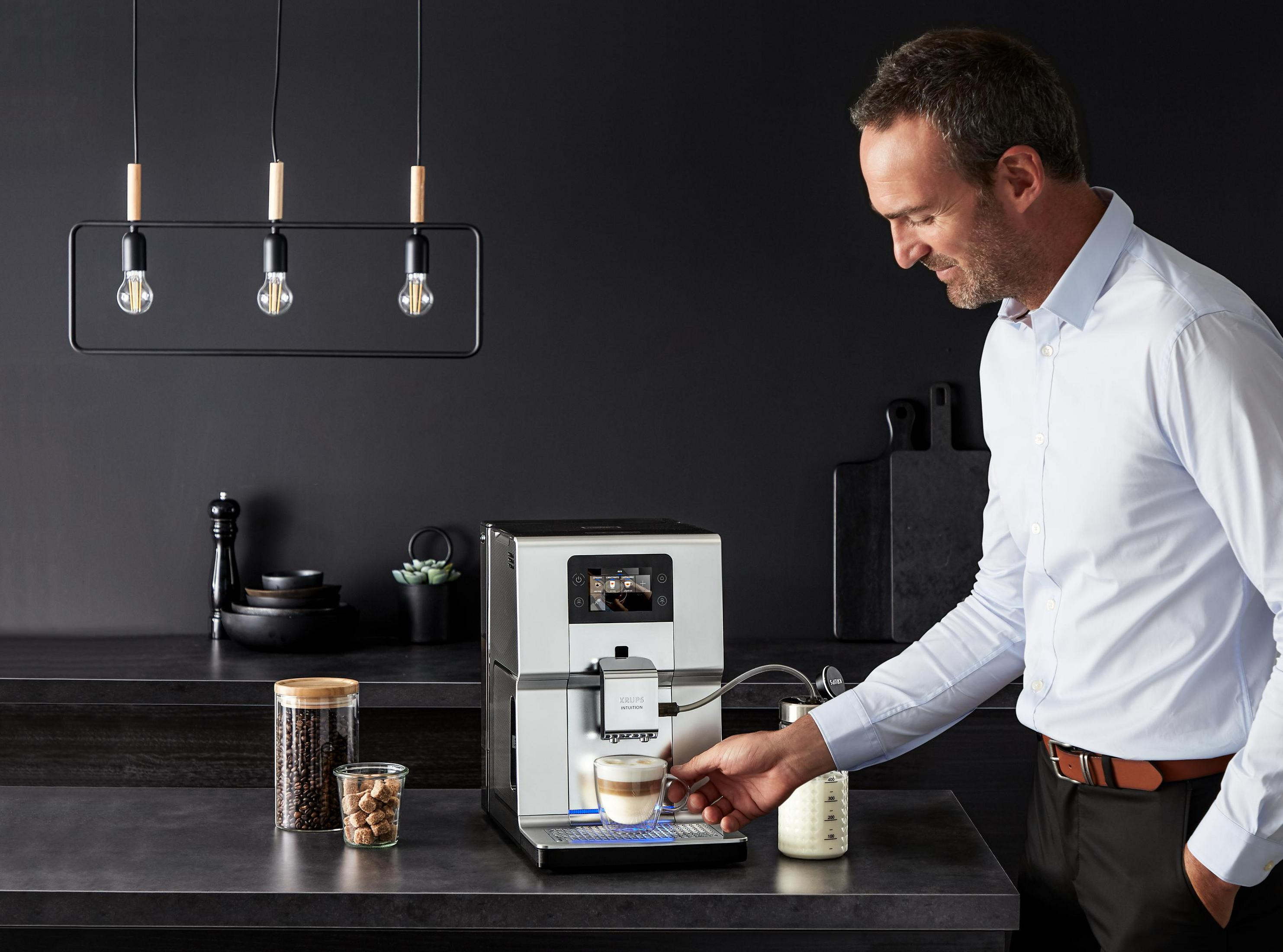 Mężczyzna obsługujący srebrno-czarny ekspres do kawy, nalewający latte do filiżanki. Ziarna kawy i cukier.