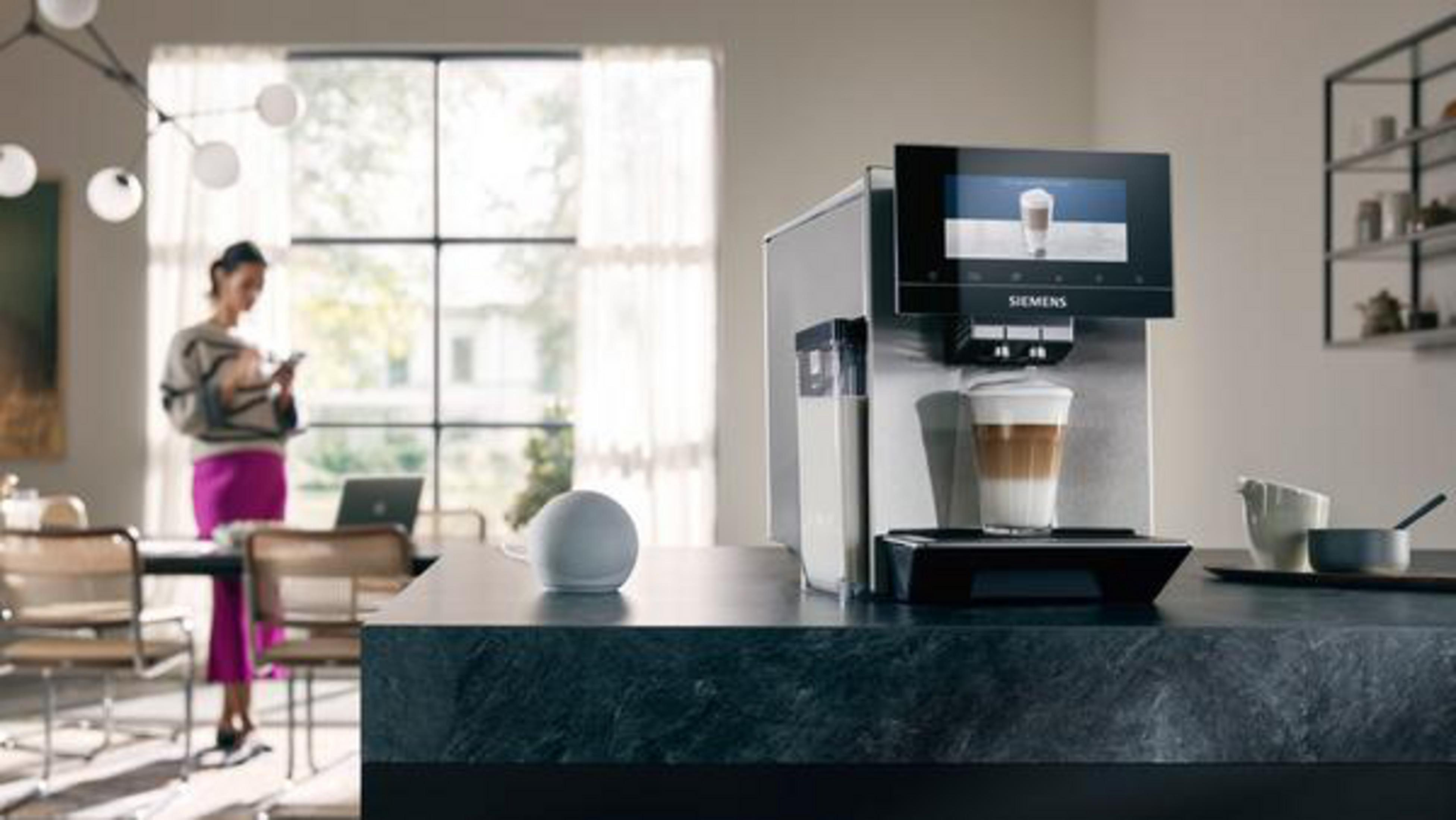 Kücheninnenraum mit Kaffeemaschine und Frau am Telefon. Kaffeetasse und Smart Speaker auf der Theke.