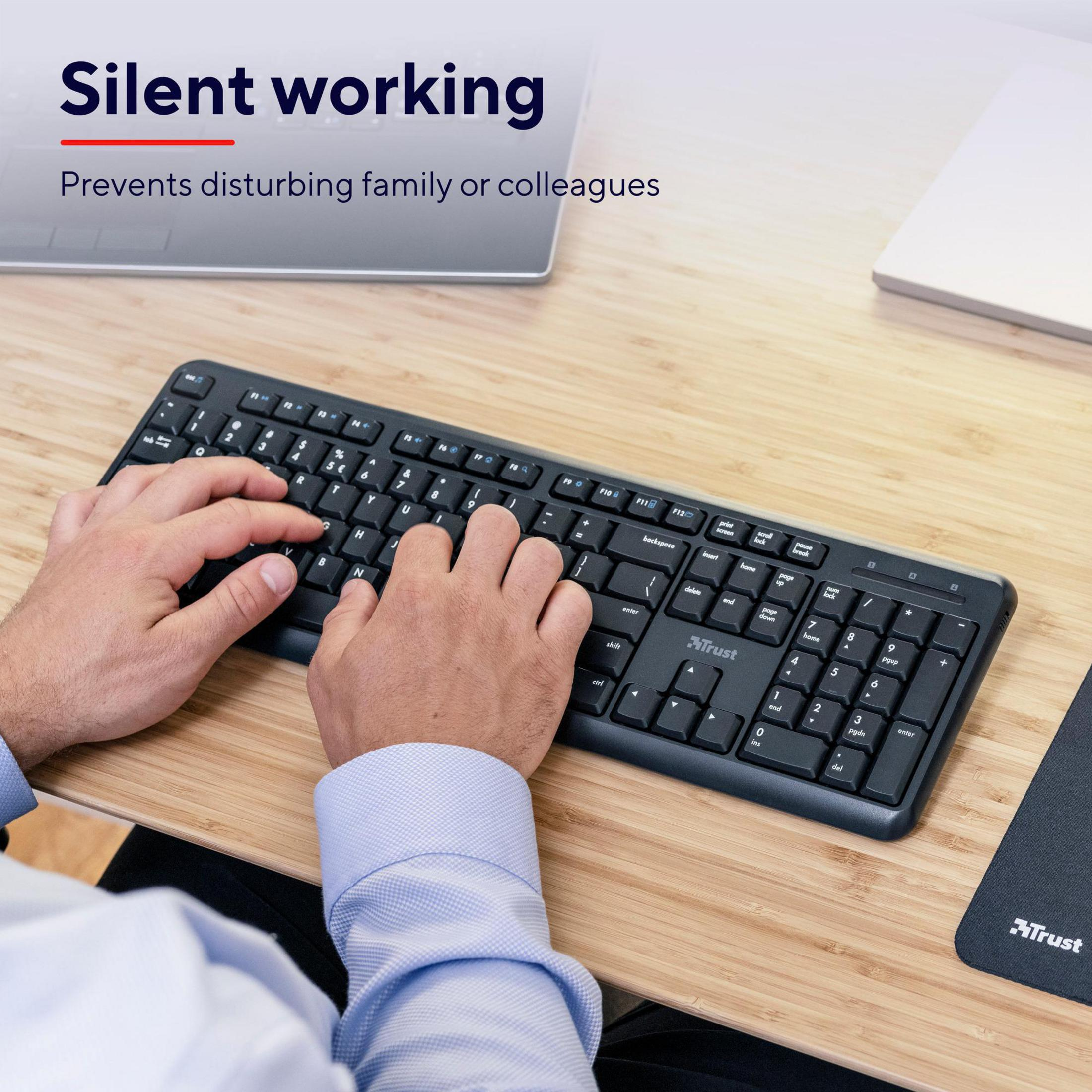 Ręce piszące na czarnej klawiaturze Trust. Klawiatura leży na drewnianym stole.