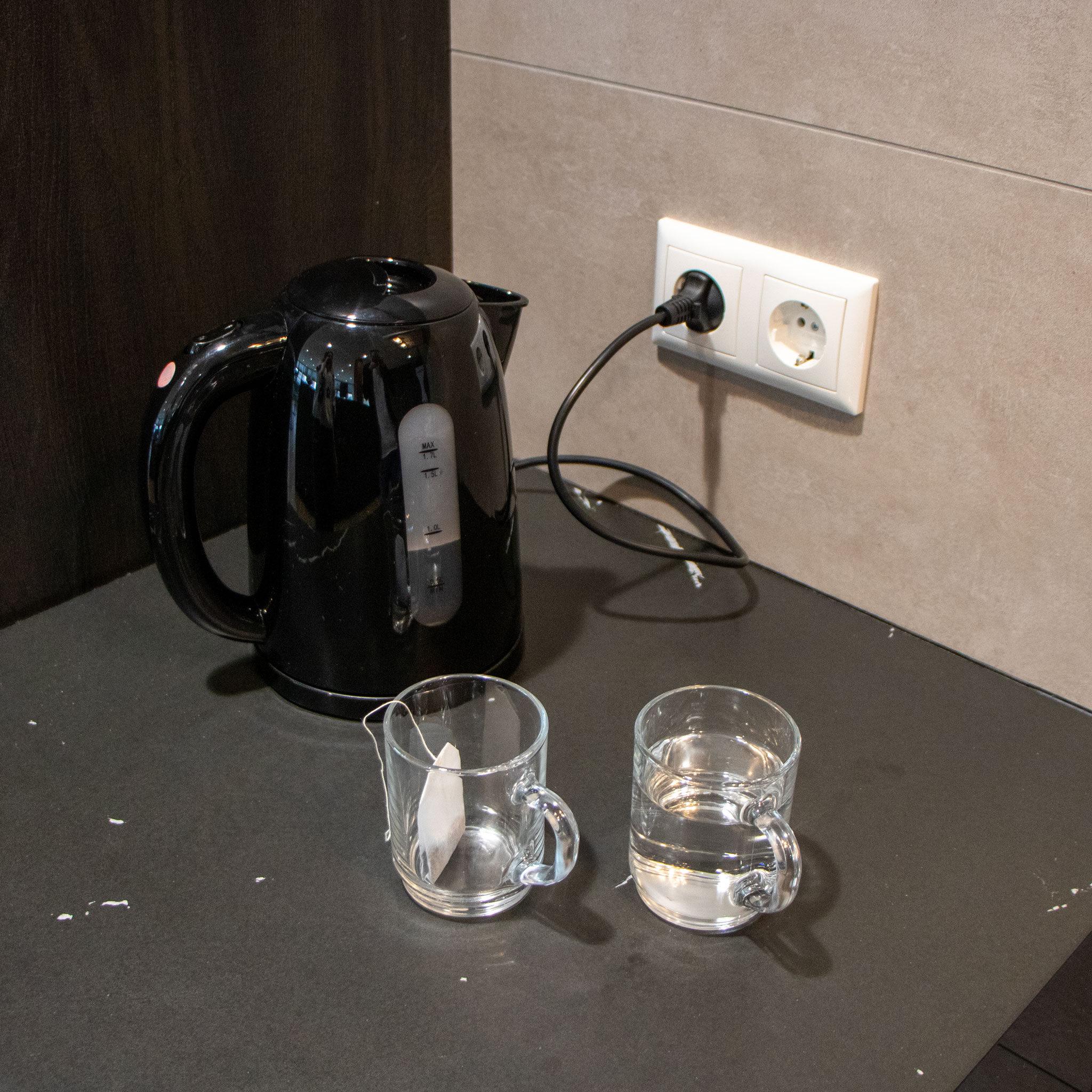 Czarny czajnik elektryczny i dwie przezroczyste szklanki z wodą. Czajnik jest podłączony do gniazdka ściennego.
