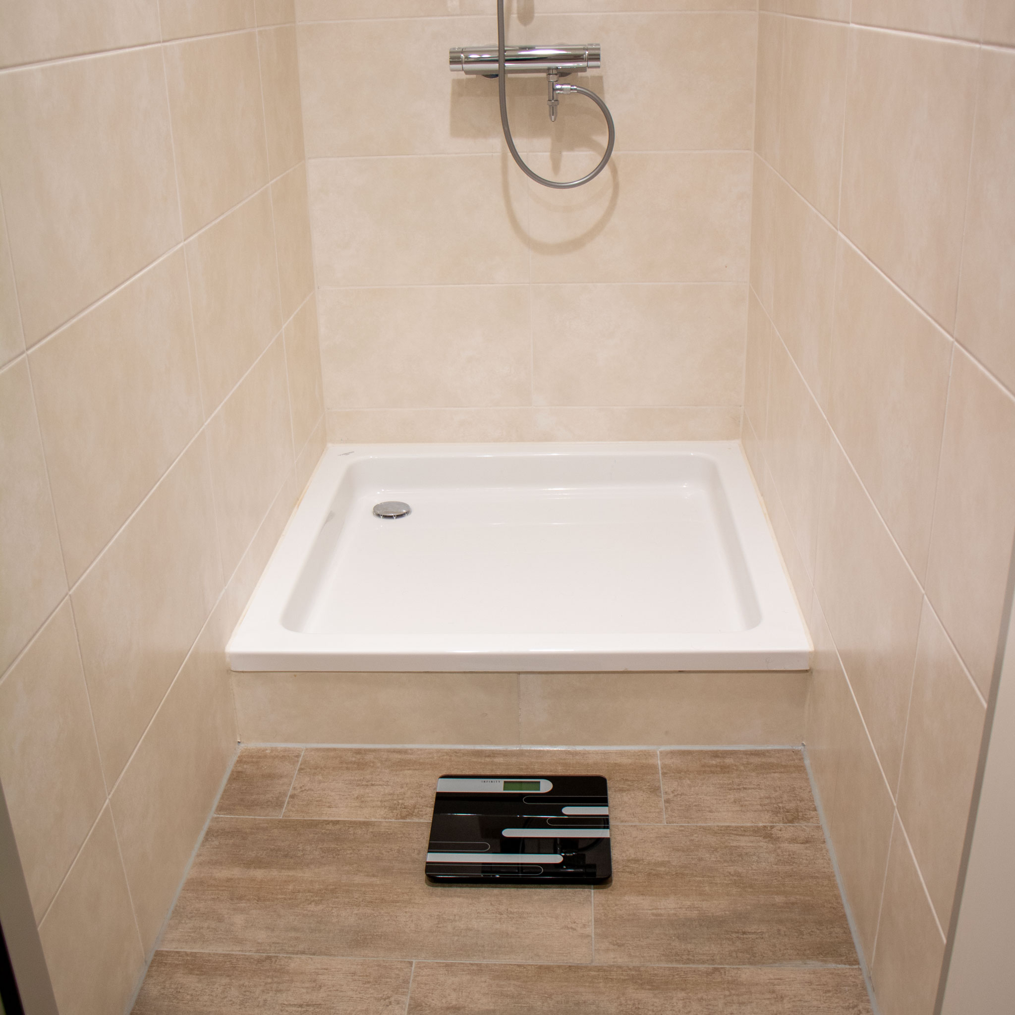 Obszar prysznica w łazience. Biały brodzik i beżowe kafelki na ścianach. Waga na podłodze.